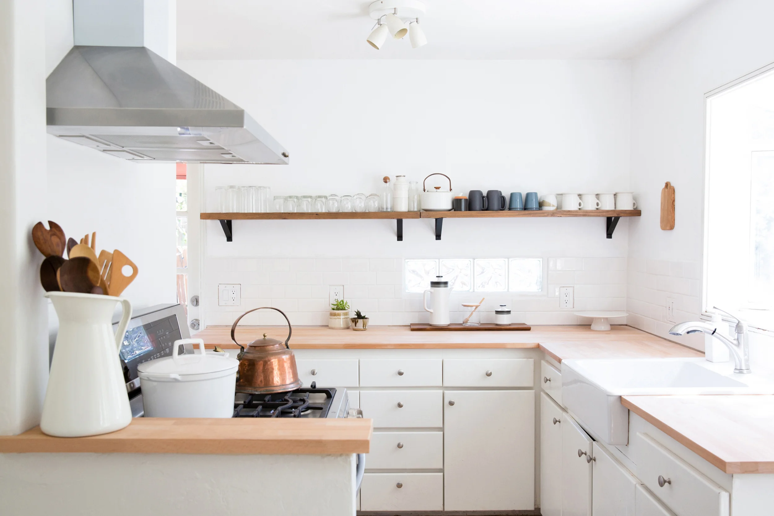 Cocina moderna con gabinetes blancos, encimera de madera, una estufa y una alzada con utensilios de cocina de madera. Estantes con vasos y tazas, lámpara de techo y ventana que deja entrar luz natural.