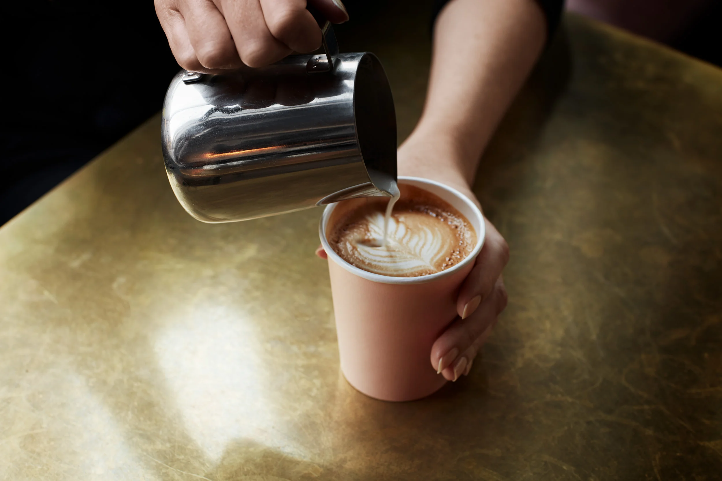 Una mano sta versando latte in un cappuccino in una tazza di carta rosa su un tavolo di legno.