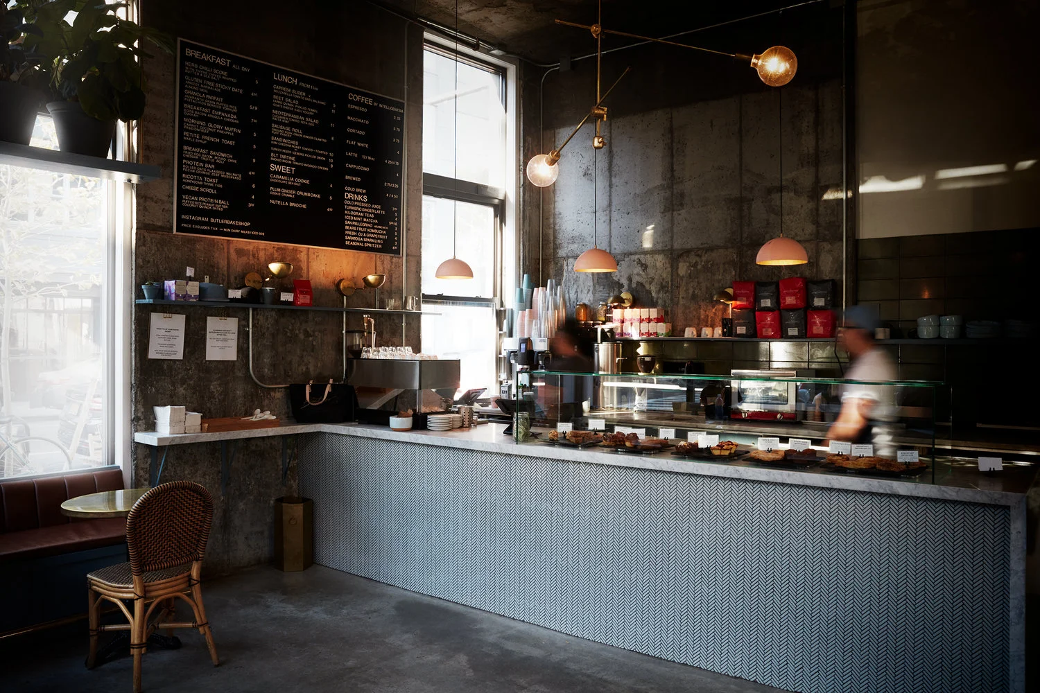Interno di un caffè con pareti di cemento, illuminazione calda con lampade appese, bancone con vassoi di dolci e pasticcini, menu sul muro, zona bar con macchina per caffè e scaffali con scatole di thè e tisane.