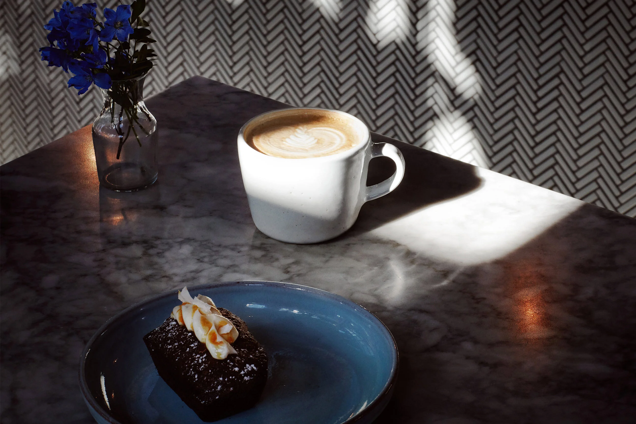 Tazza di caffè con latte e un dolce al cioccolato con panna su un piatto blu, su tavolo di marmo, luce naturale e sfondo con pattern geometrico.