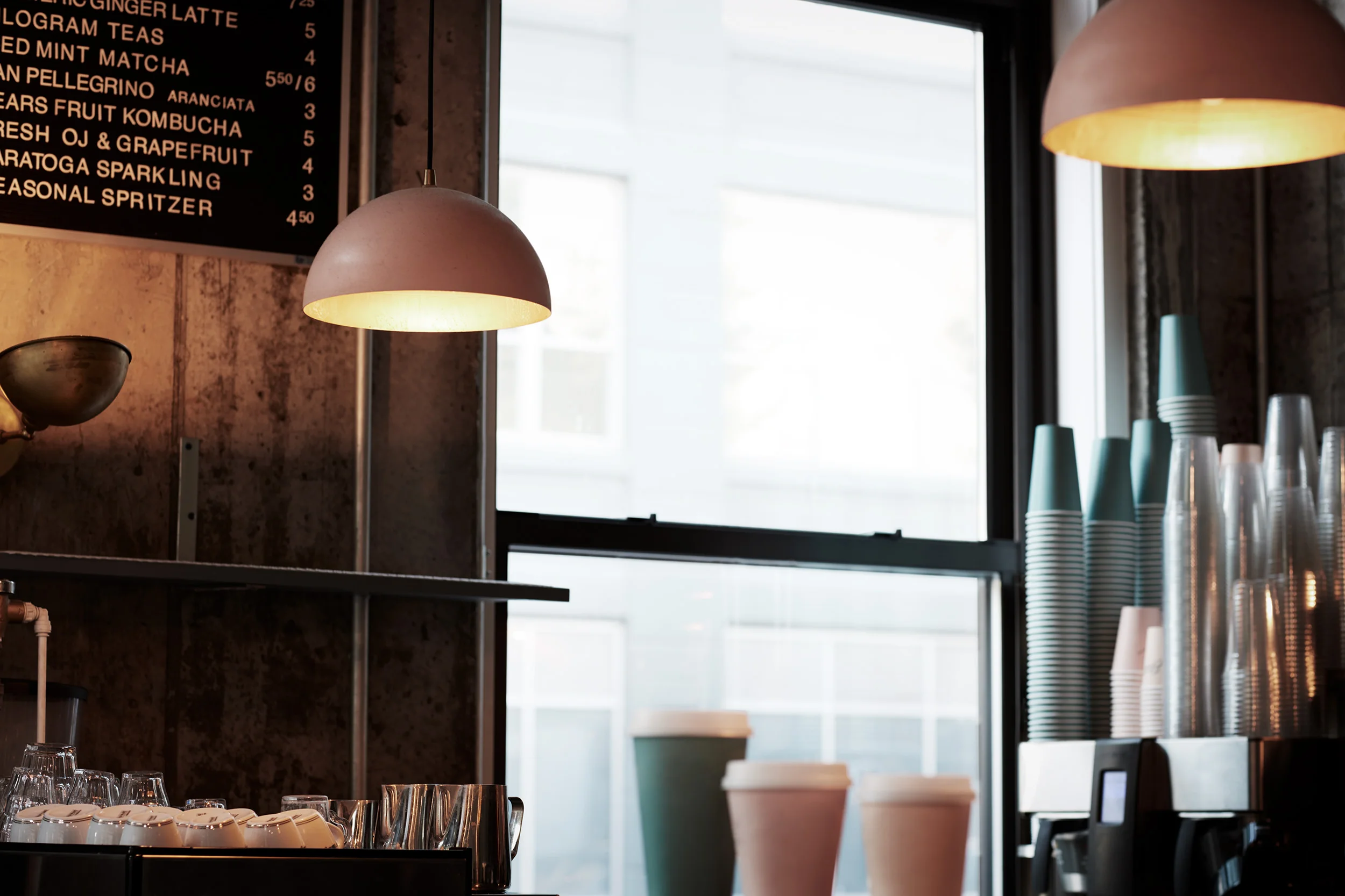 Interno di un bar con lampade appese, menu scritto a parete, bicchieri e contenitori di carta, e una grande finestra