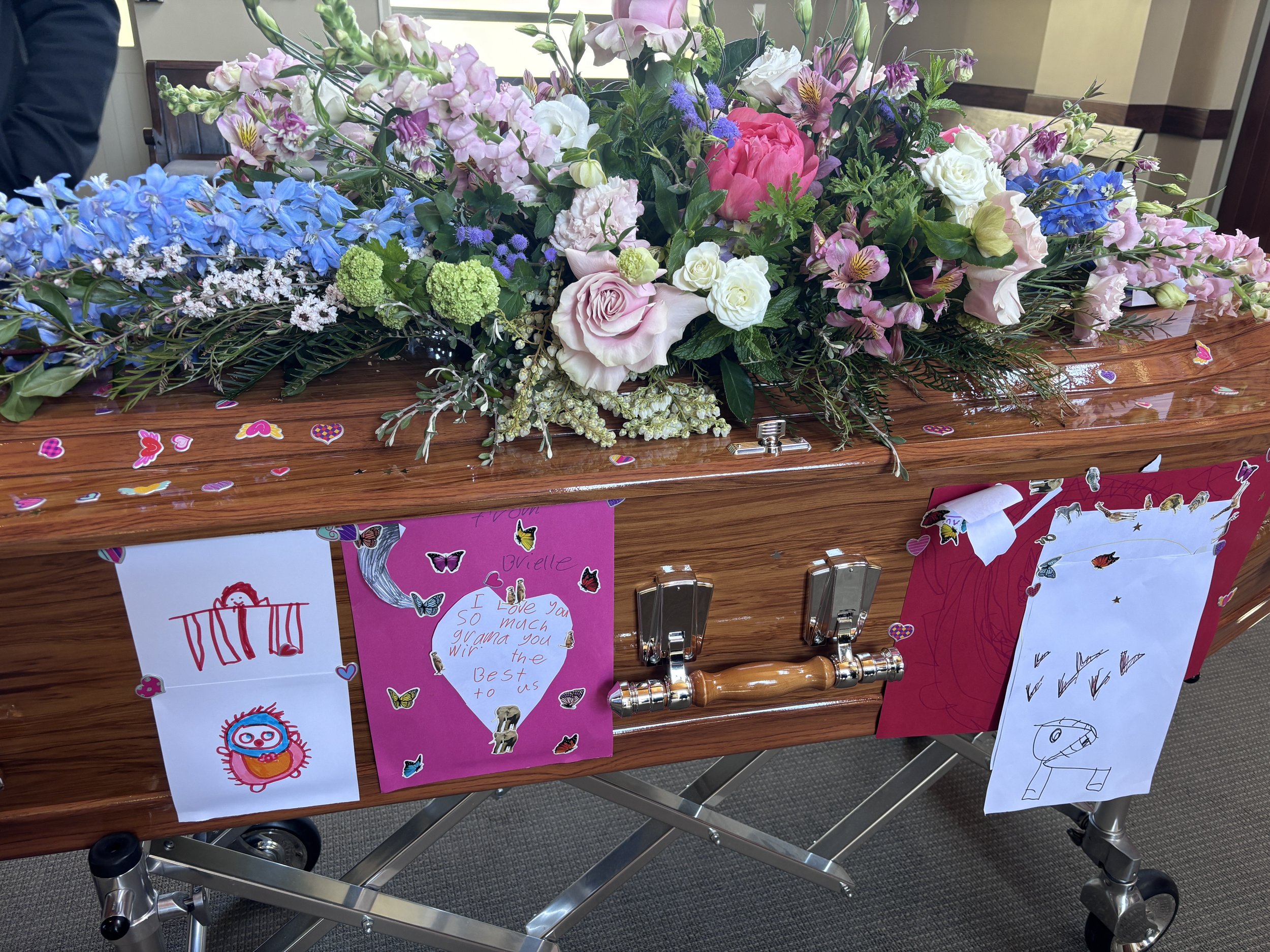 A wooden casket decorated with pastel-colored flowers and children's drawings attached to its side, with small sticker hearts scattered on the surface.
