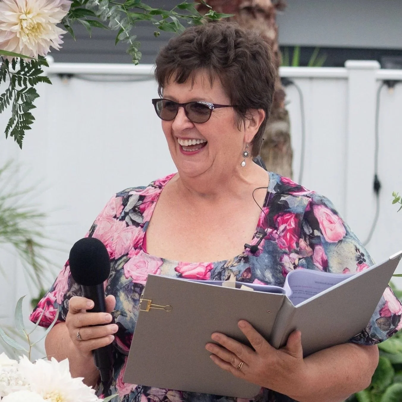 Celebrant Debbie laughing in a ceremony, holding her microphone and folder.
