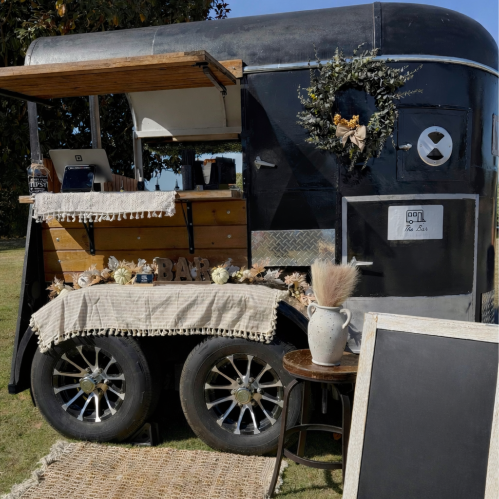 A vintage black food truck decorated with a wreath, a sign labeled 'The Bar,' and various decorations including a vase with pampas grass on a small table in front. It has a wooden serving counter with a lace cloth, pumpkins, and other decorative items. There is a laptop and a bottled drink on the counter, with a decorative chalkboard sign and a chalkboard menu nearby.