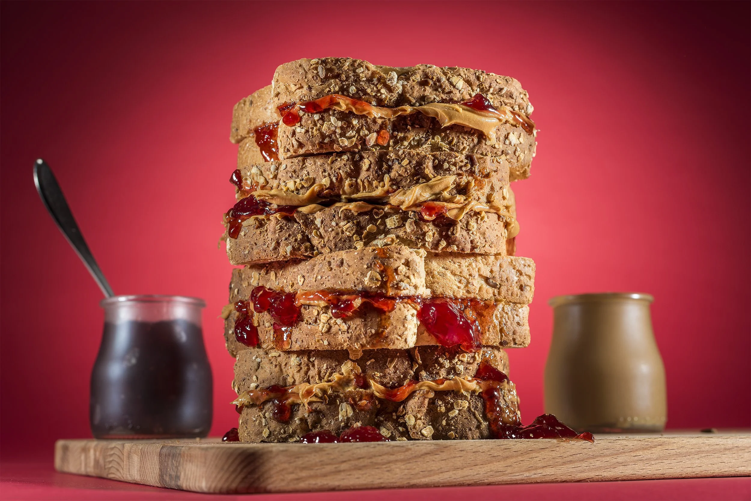 A tall stack of whole grain sandwiches filled with peanut butter and strawberry jelly with jars in the background