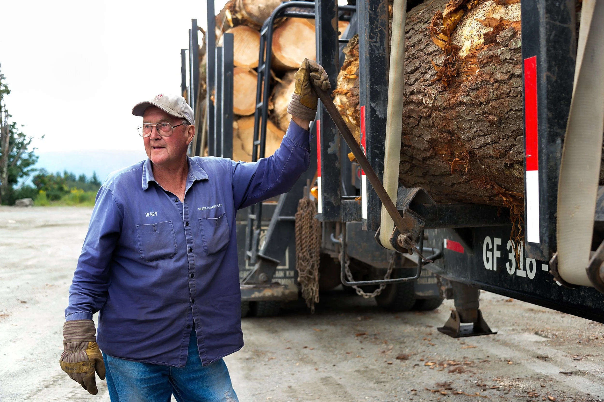 Older man in work clothes leaning on logging truck loaded with logs