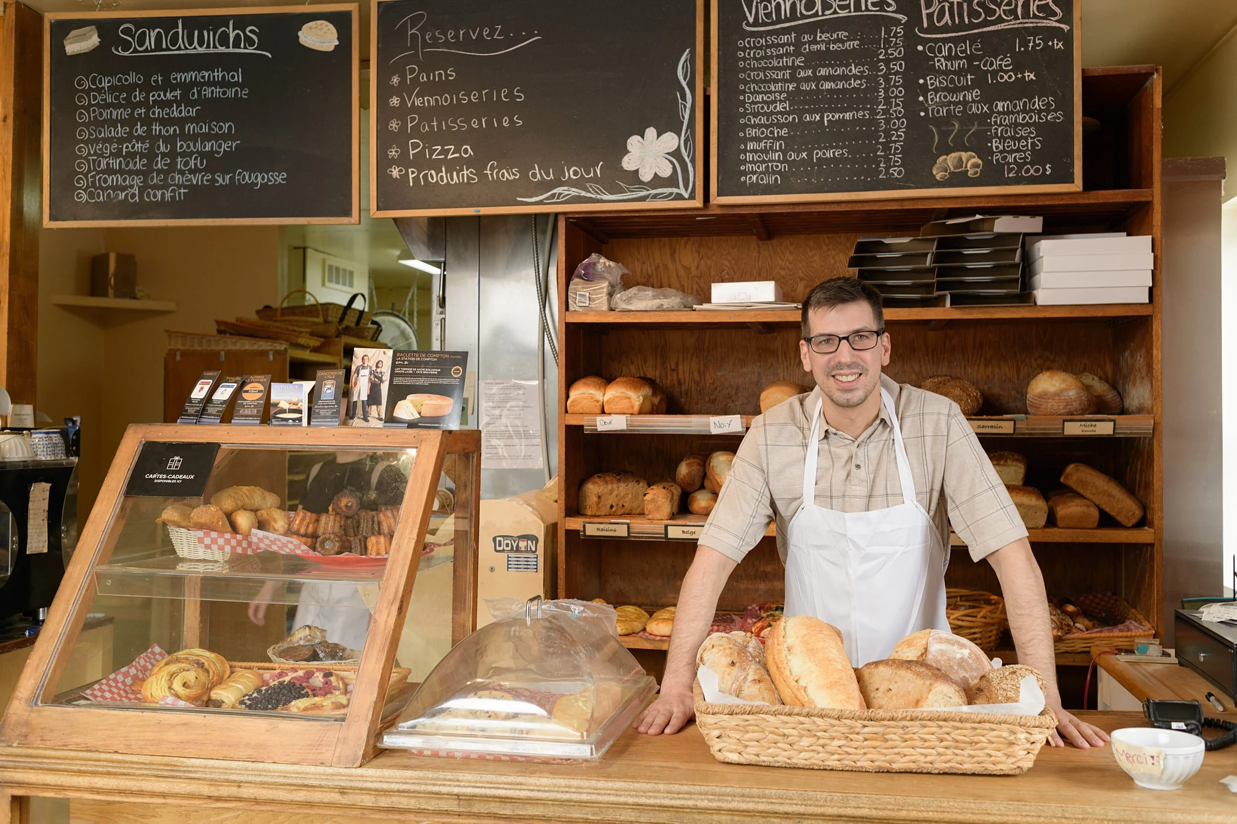 Male baker leaning on a bakery display counter with bread loaves in baskets and handwritten French chalkboard menus on the wall behind him.