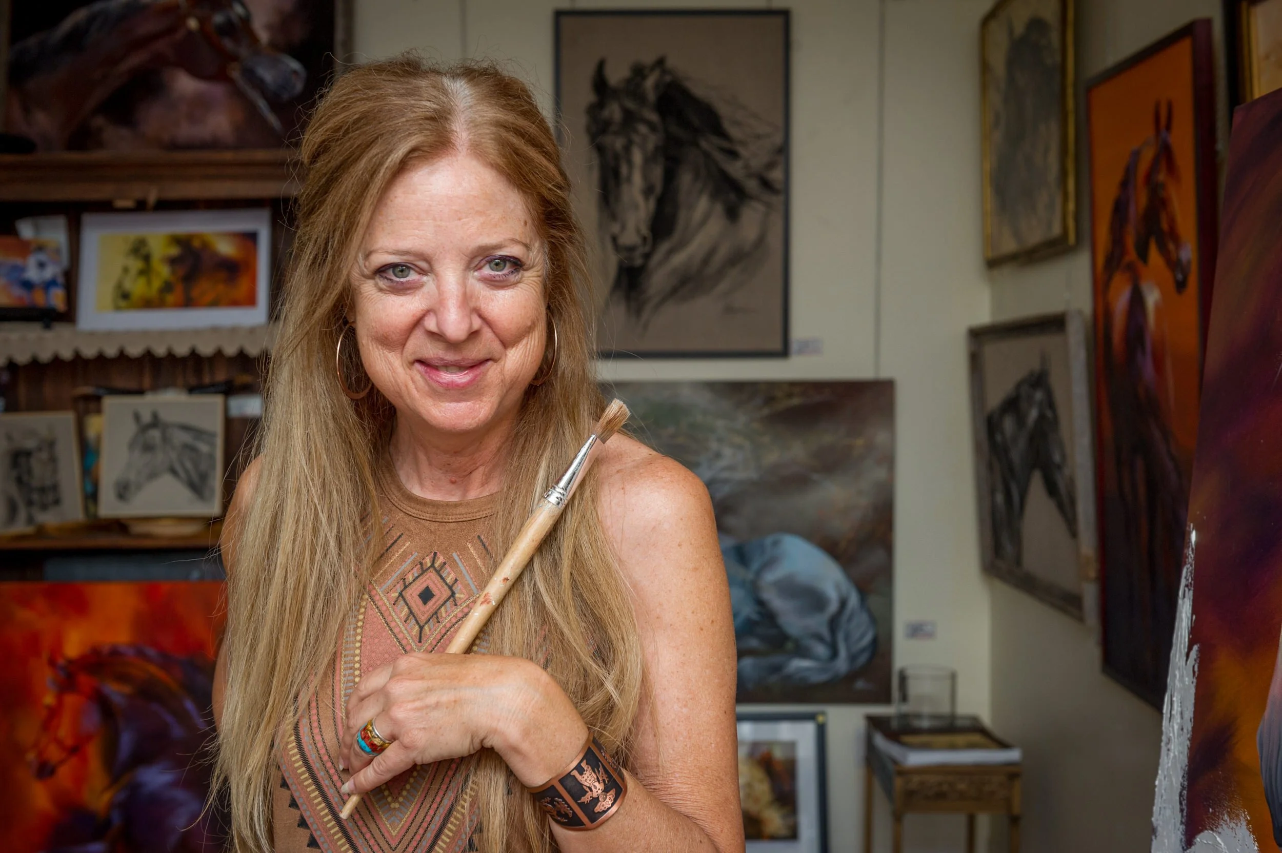 Johanne Blaquière Smiling and holding paintbrush in a studio filled with horse paintings