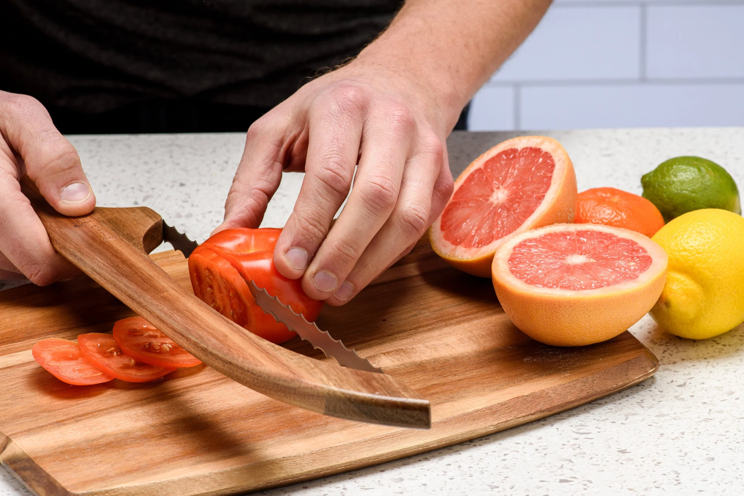 Hands using the Green Ara knife to slice a tomato on a wooden board