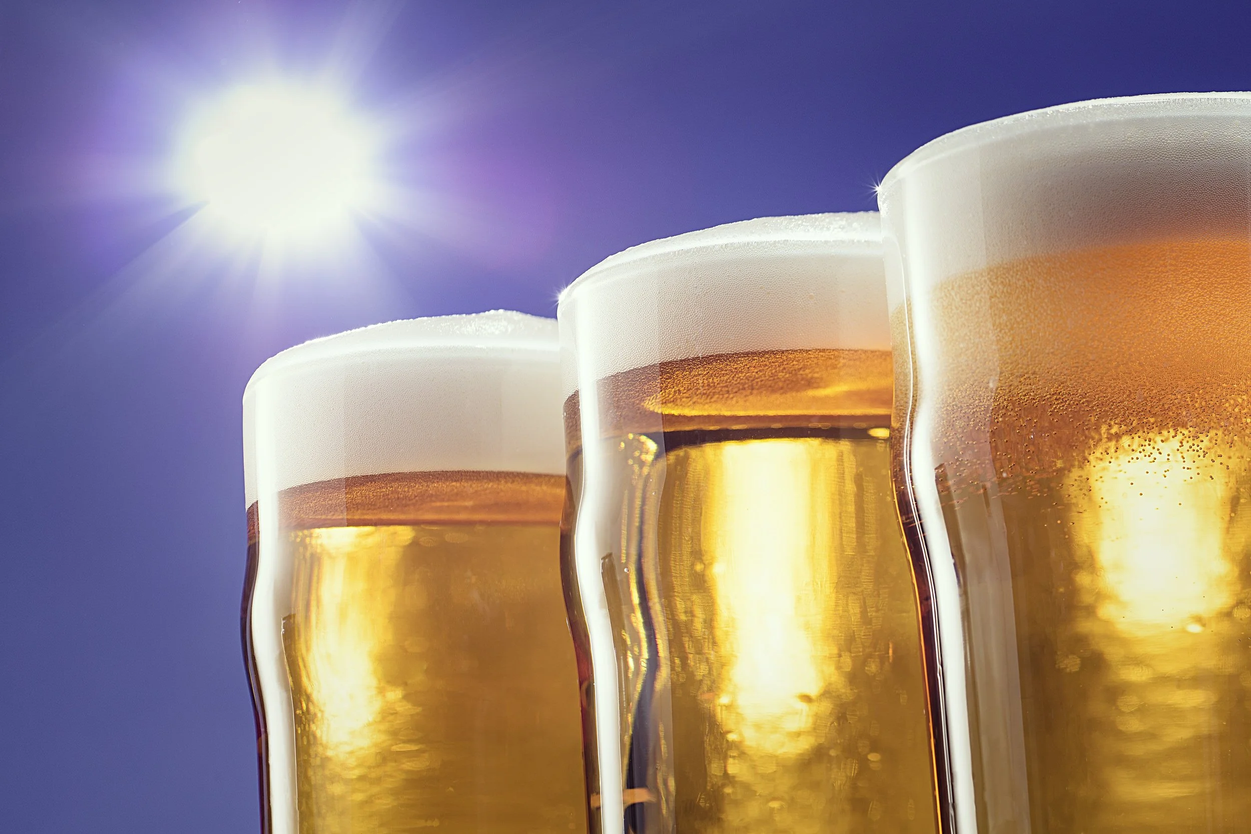 Three golden beers in glasses with foamy heads against a sunny sky