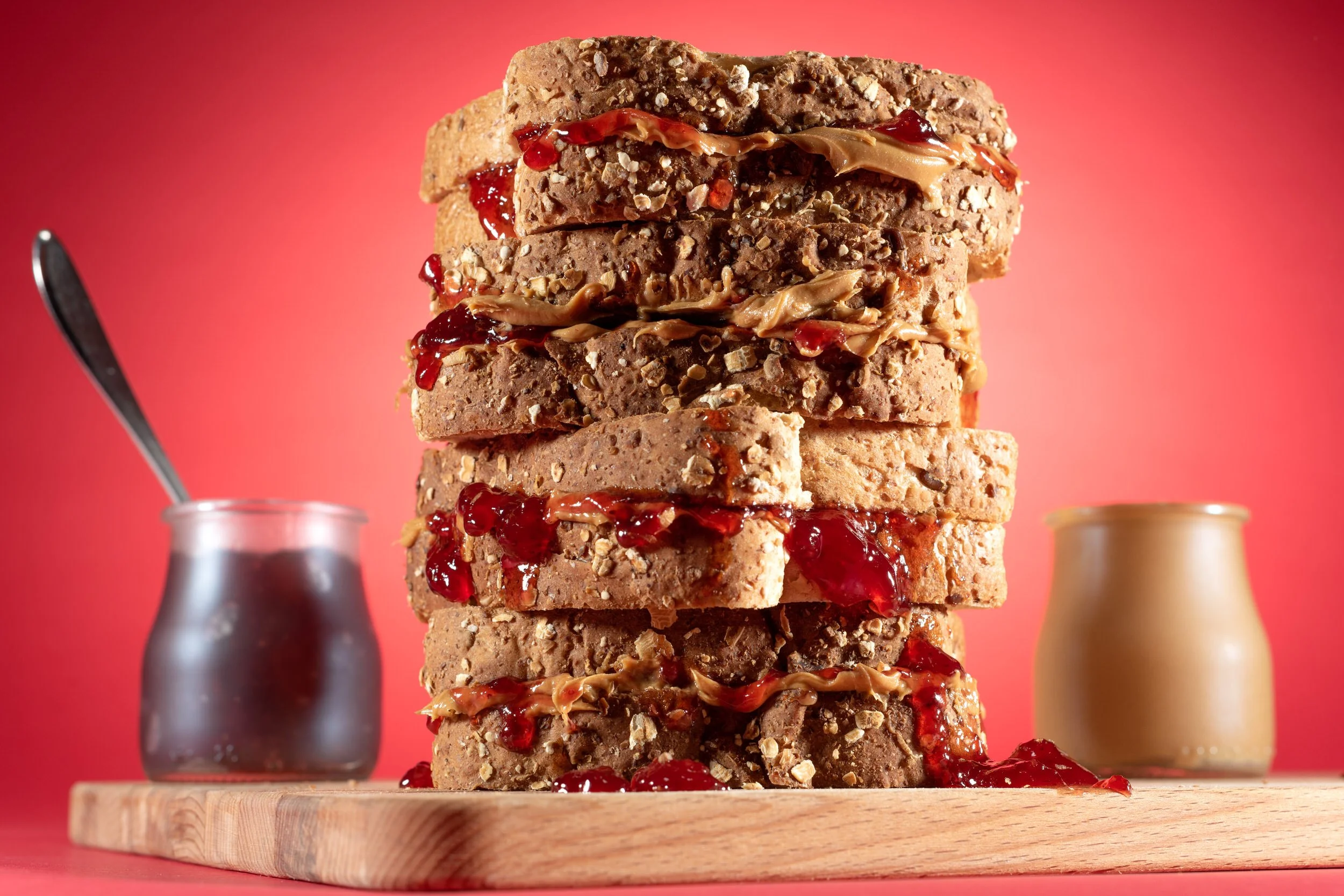 A tall stack of whole grain sandwiches filled with peanut butter and strawberry jelly with jars in the background