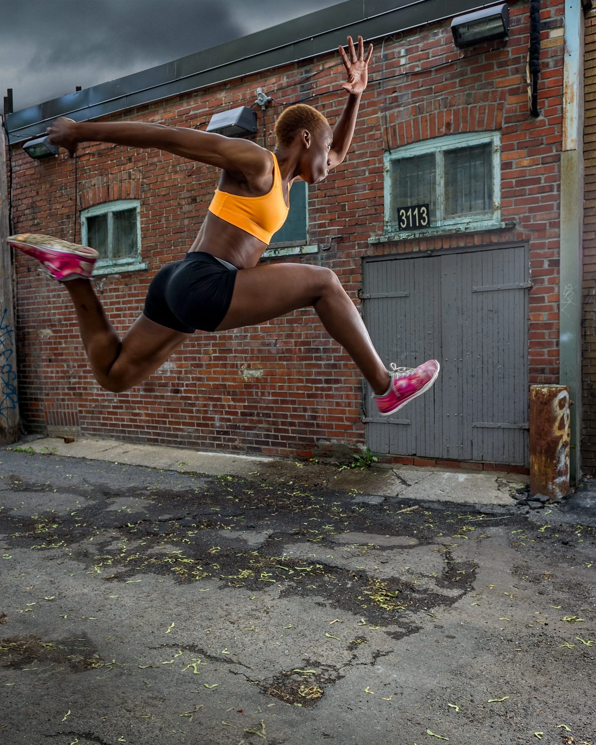 Female athlete mid-jump in an urban alley wearing an orange sports bra and black shorts, with a dark stormy sky and red brick building behind her.