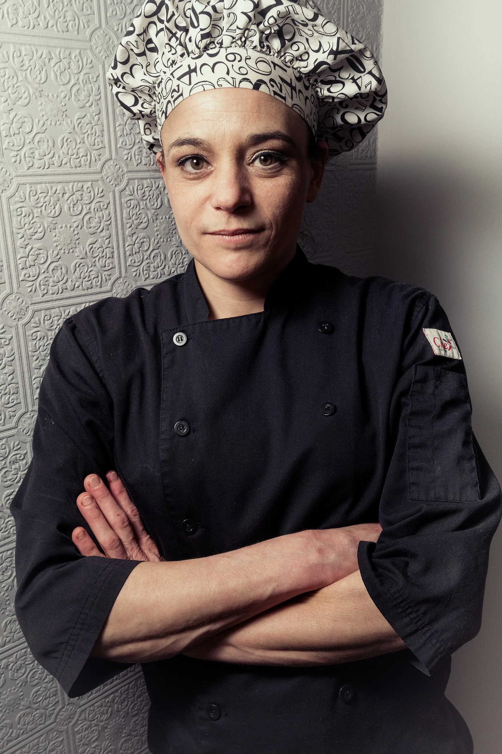 Female pastry chef in black uniform with arms crossed, looking at camera with a composed, confident expression against a white embossed wall.