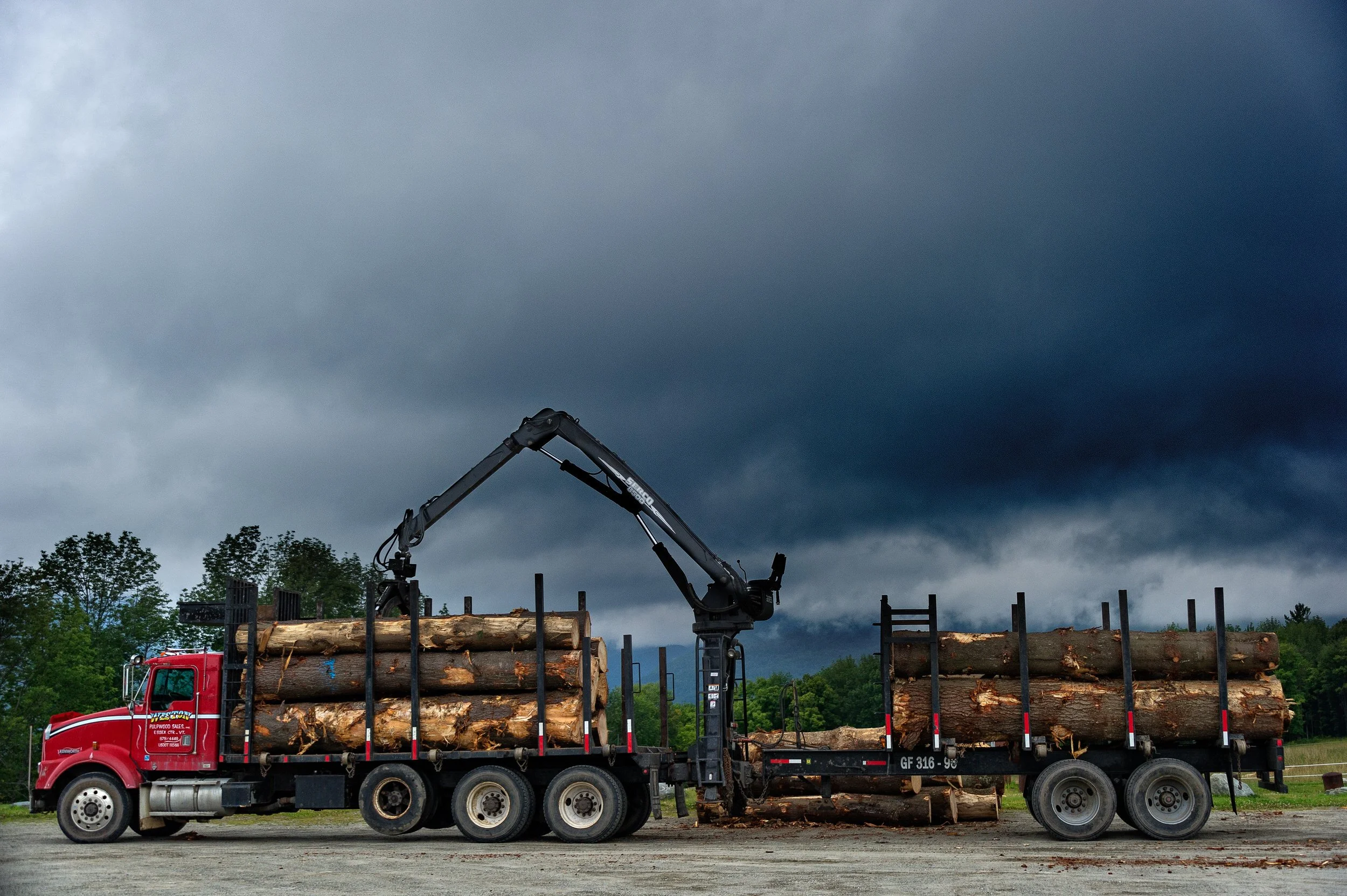 Red logging truck loaded with logs parked under dark storm clouds