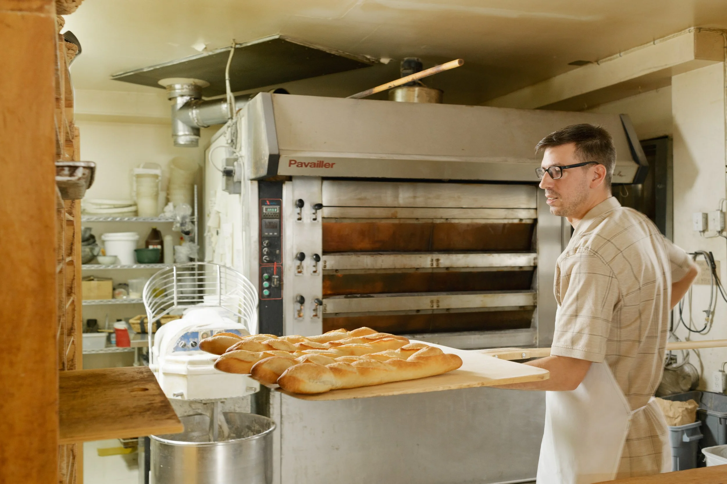 Baker removing freshly baked baguettes from a commercial deck oven using a long wooden bread peel.