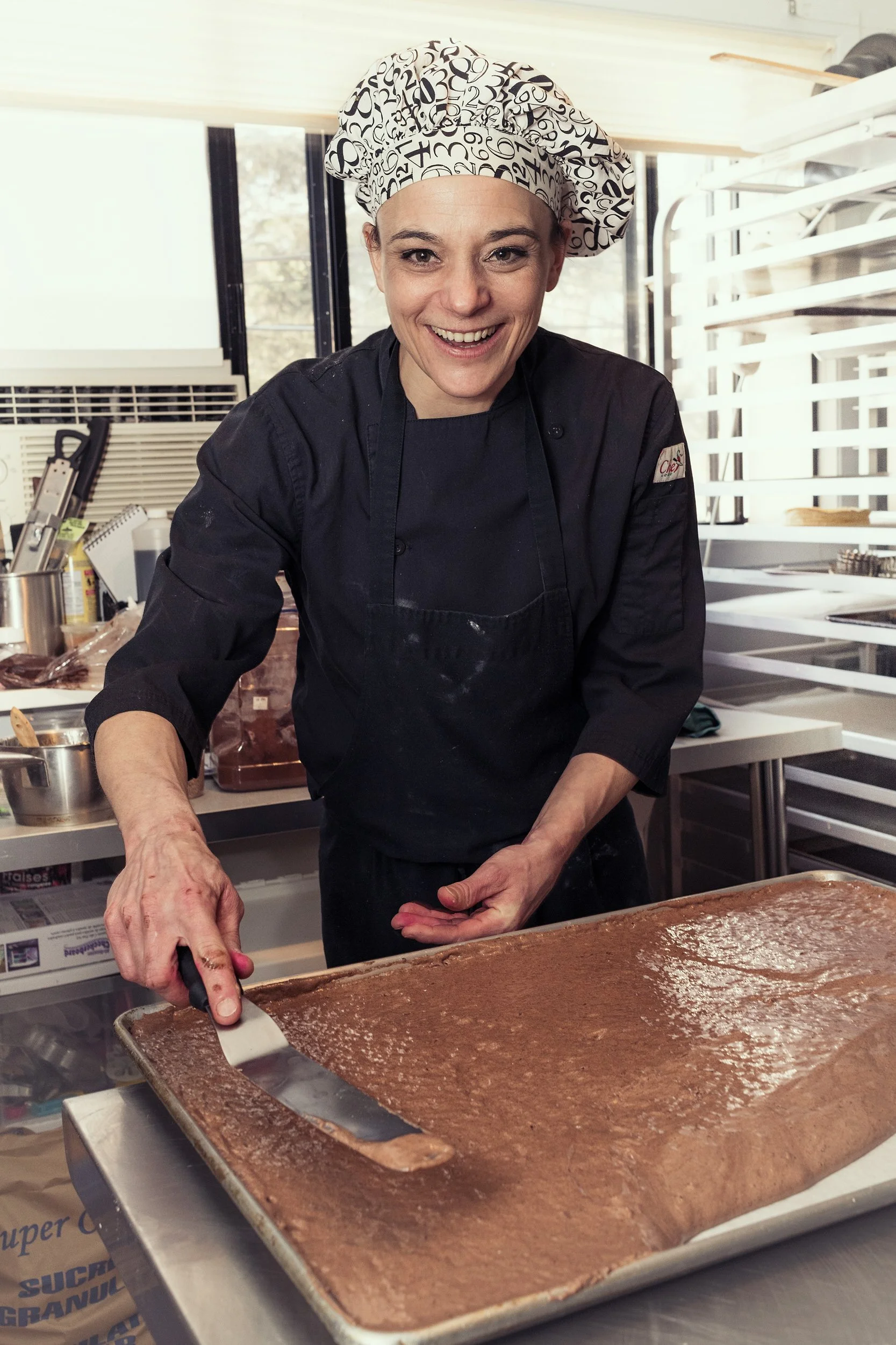 Female pastry chef in black uniform spreading chocolate batter on a large baking sheet with an offset spatula, smiling at the camera.