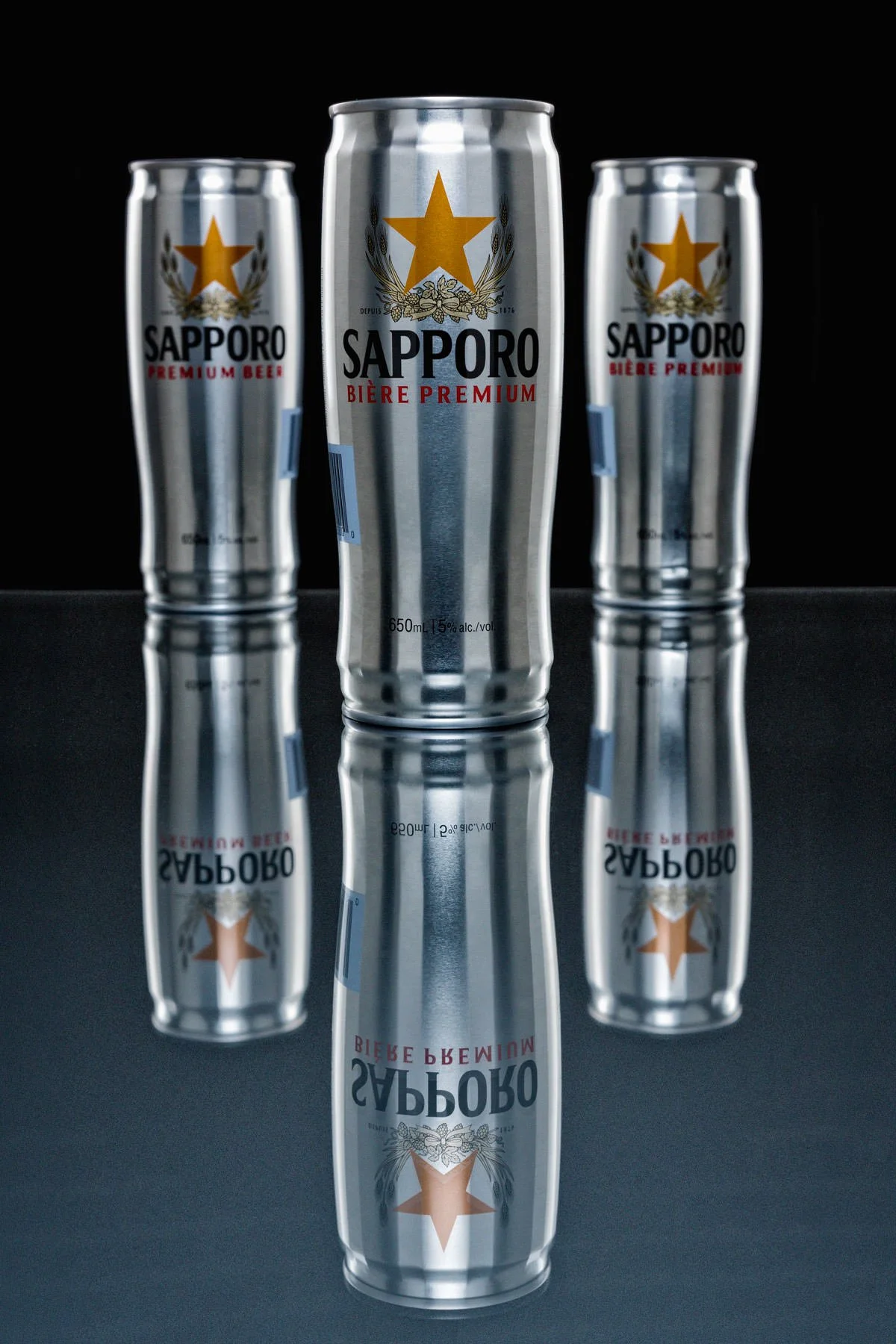 Three Sapporo beer cans standing on reflective glass with dramatic light and branded packaging