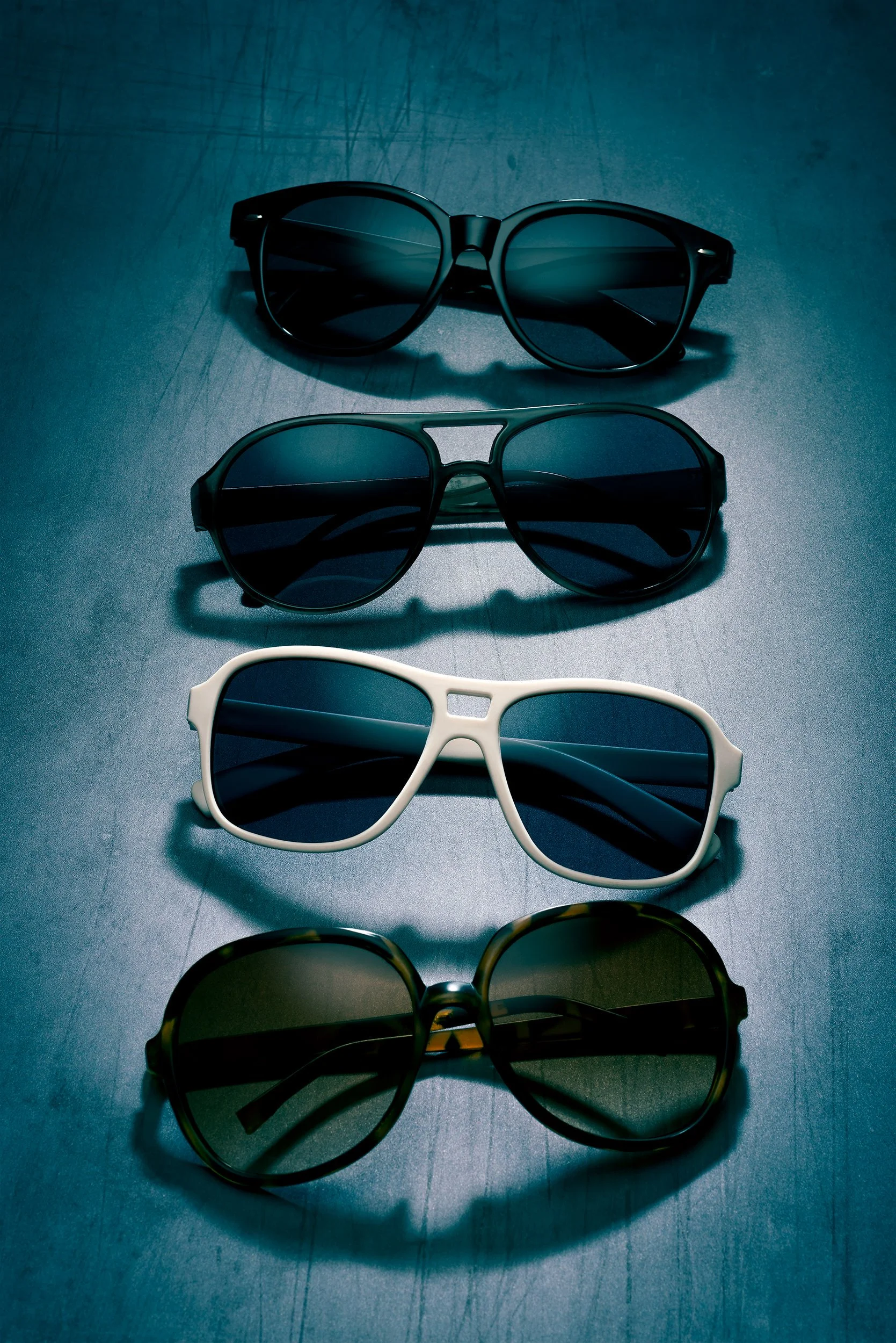 Top-down view of four pairs of sunglasses aligned in a column on a textured blue-black background