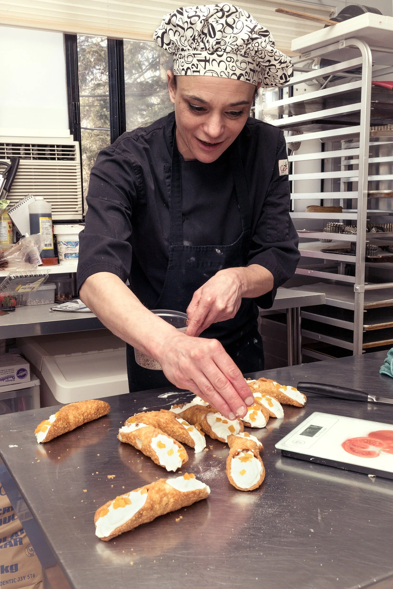 Pastry chef sprinkling garnish onto a row of cream-filled cannolis on a stainless steel counter in a professional kitchen.