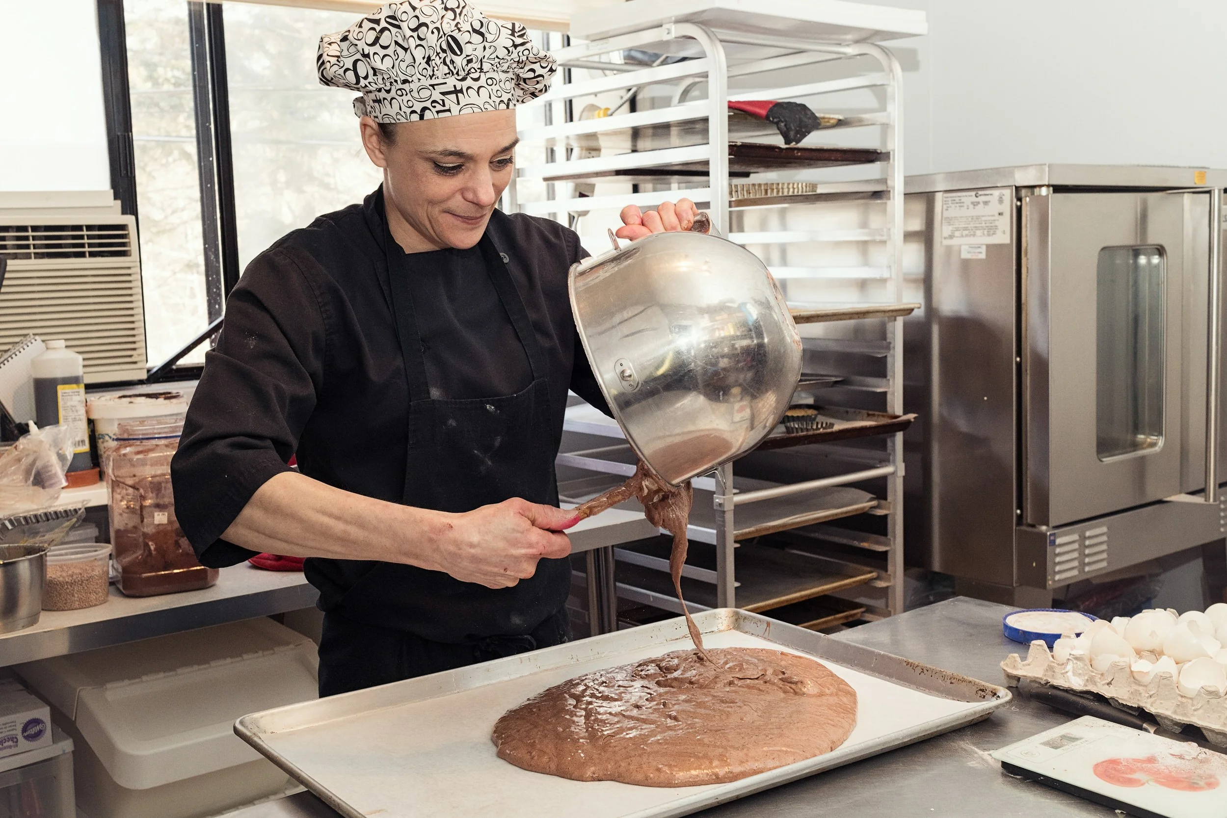 Pastry chef pouring thick chocolate batter from a stainless steel mixing bowl onto a baking sheet, using a red spatula to control the flow.