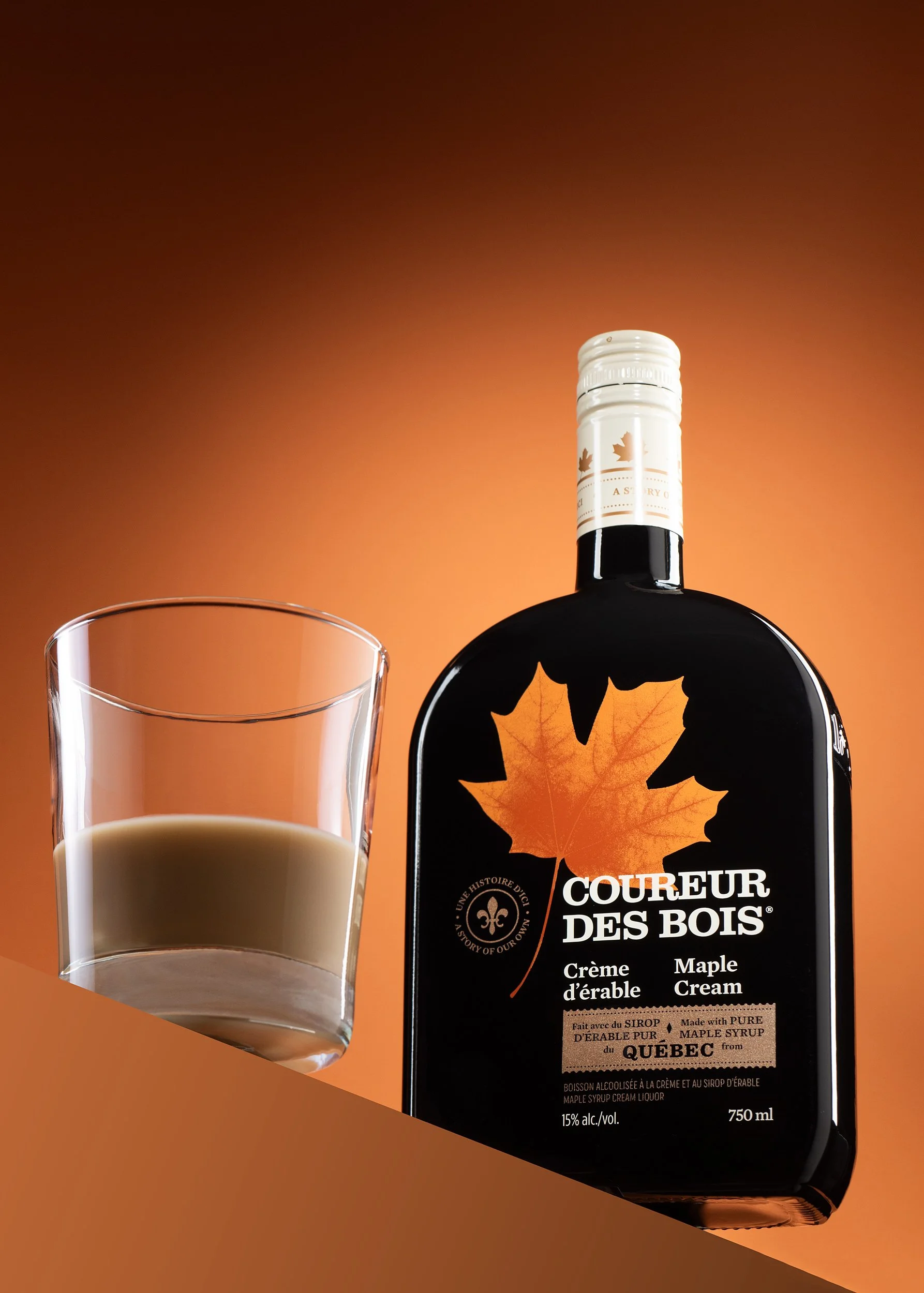Coureur des Bois maple cream liqueur bottle beside a glass of poured cream liqueur on a graduated orange background.