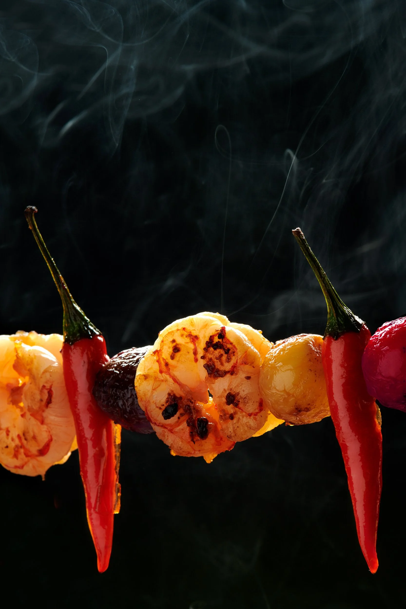 Skewer of shrimp and chili peppers against a dark smoky background with rising steam