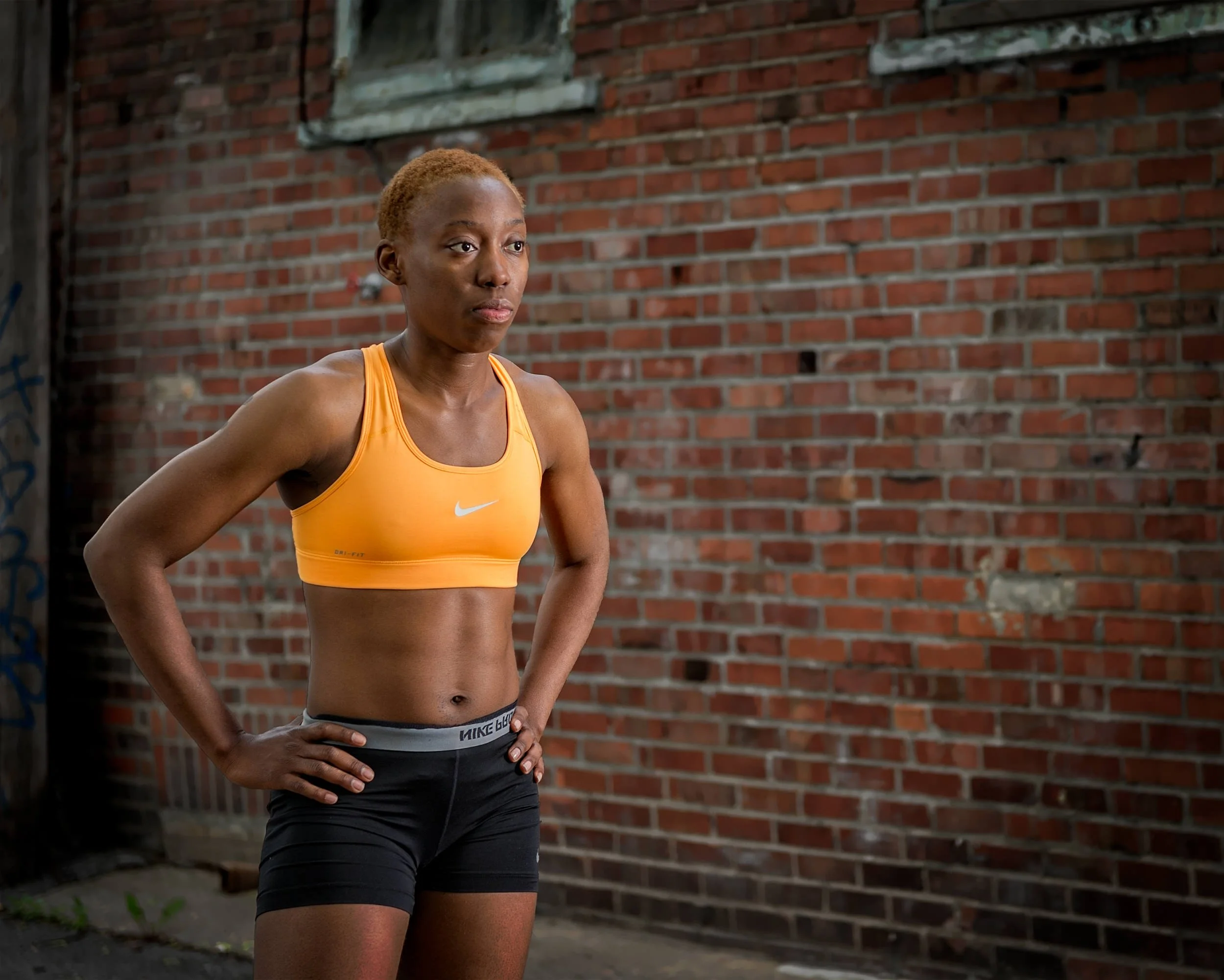 Female athlete in orange Nike sports bra and black shorts standing with hands on hips against a red brick wall, looking to the side with a composed expression.