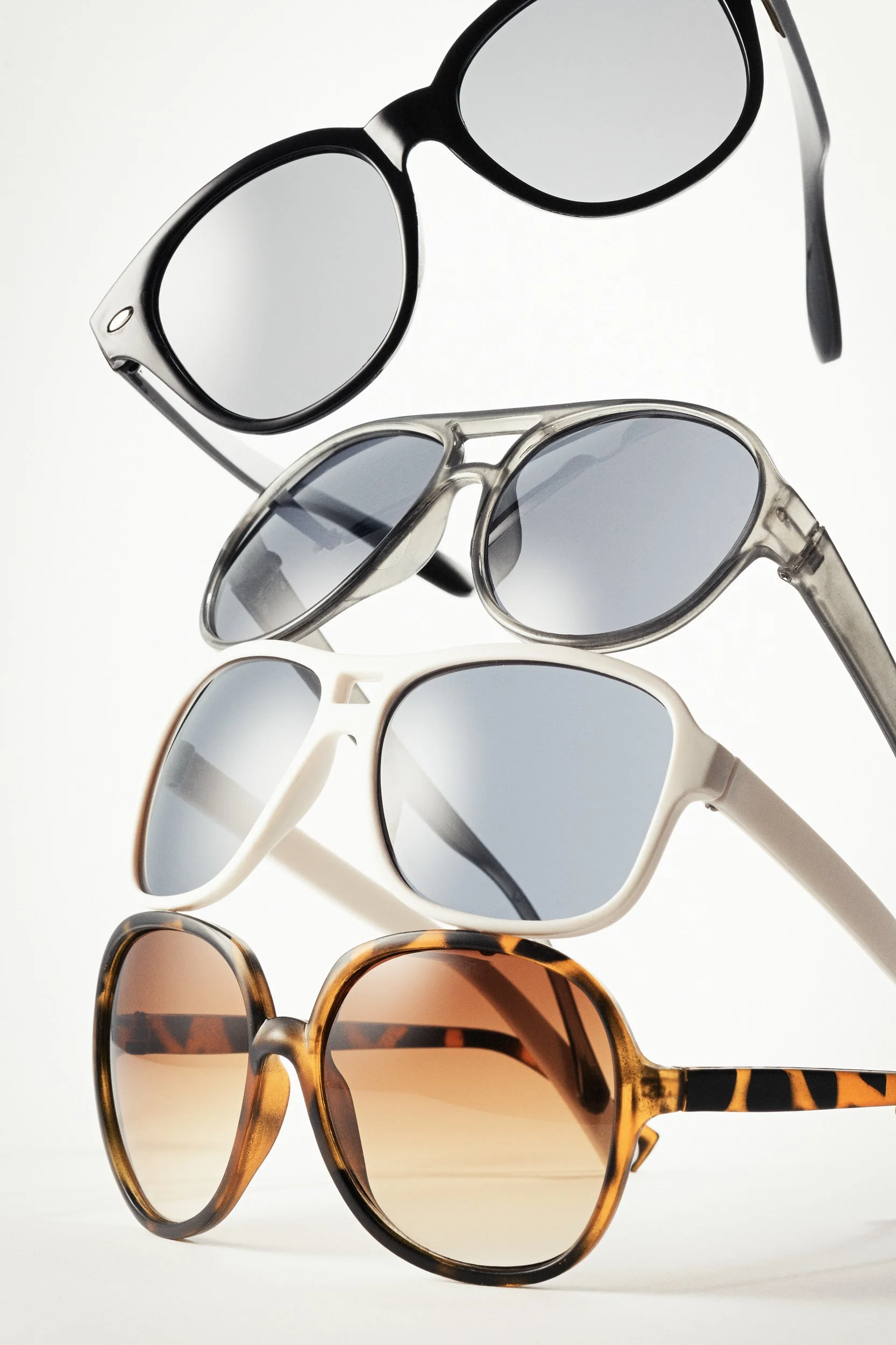 Four stacked sunglasses of various styles and colors against white background