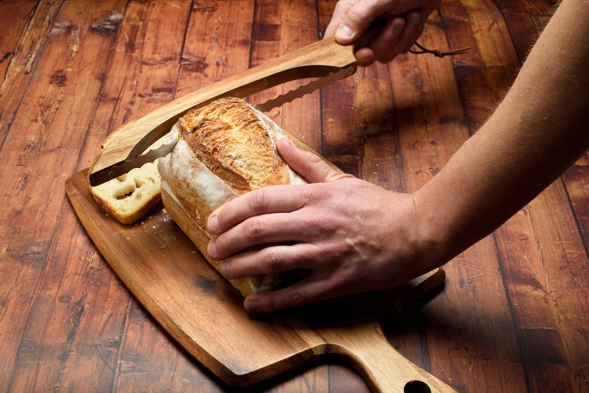 Hands using the Green Ara knife to cut through a crusty loaf on a wooden board