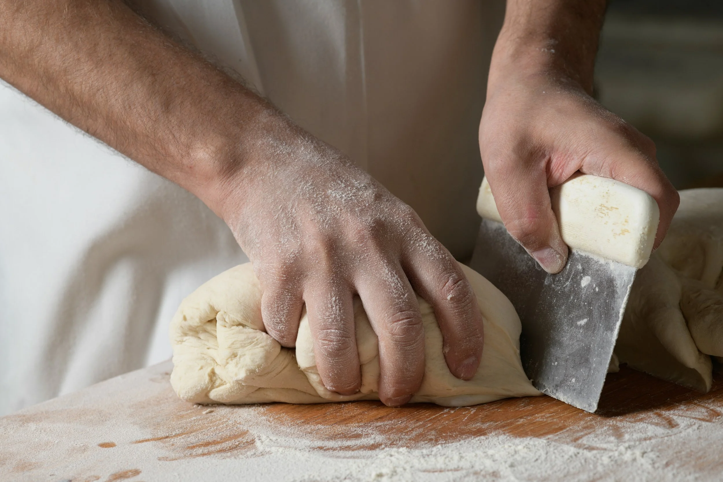 Close-up of a baker's flour-dusted hands cutting bread dough with a metal bench scraper on a floured wooden work surface.