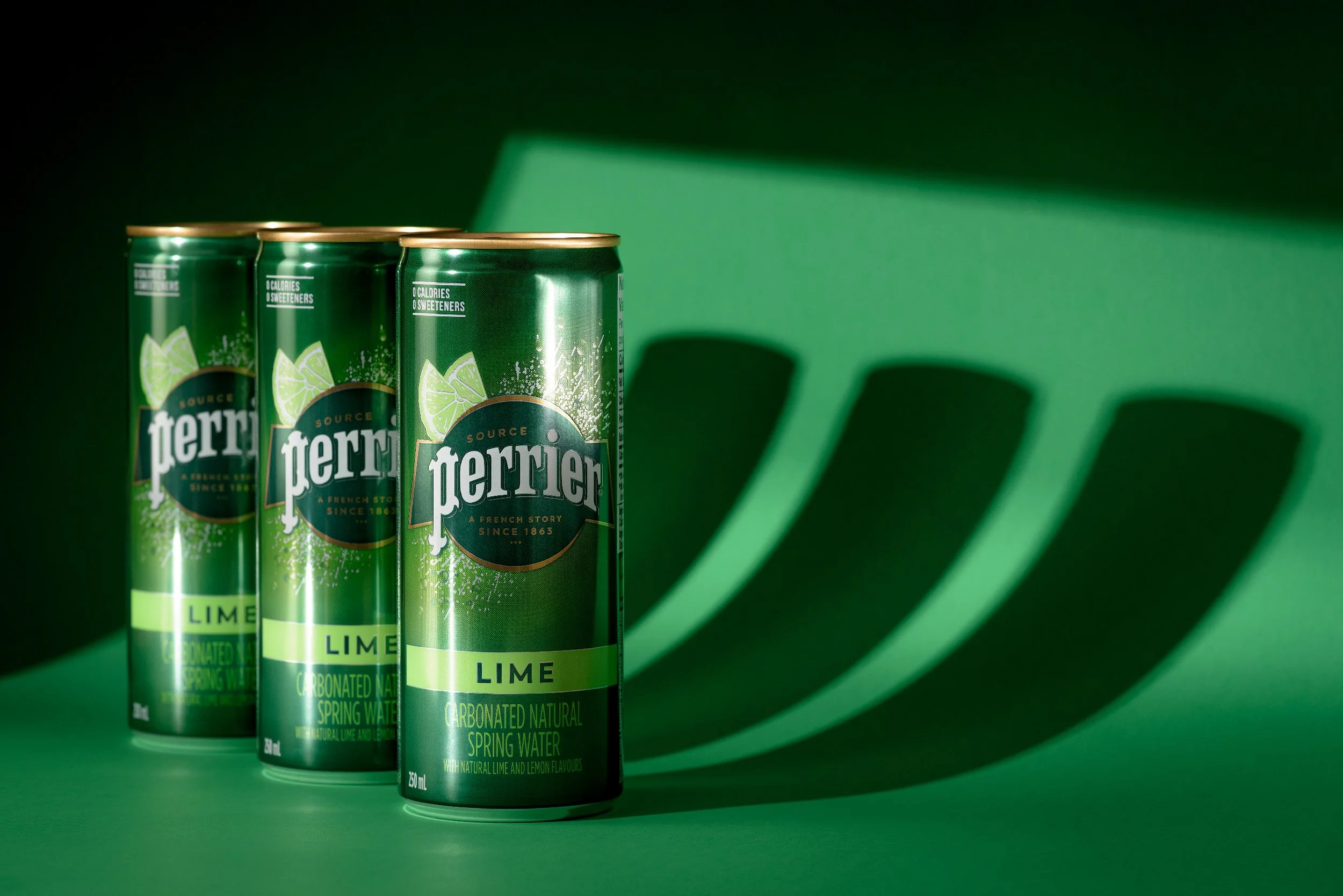 Three Perrier Lime cans casting graphic shadows on a green surface