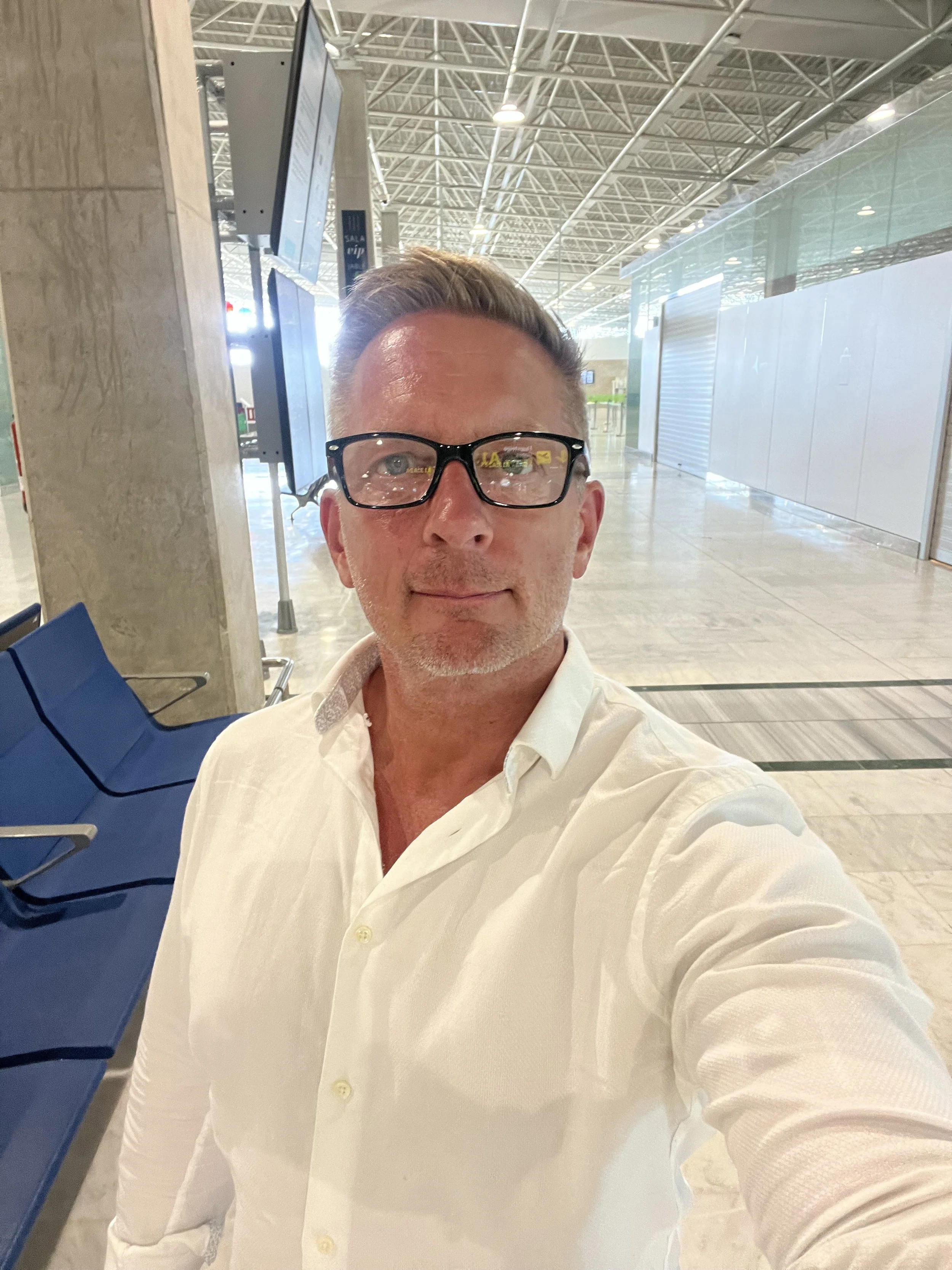A man with glasses and a white shirt taking a selfie inside an airport terminal with empty seating and high ceilings.
