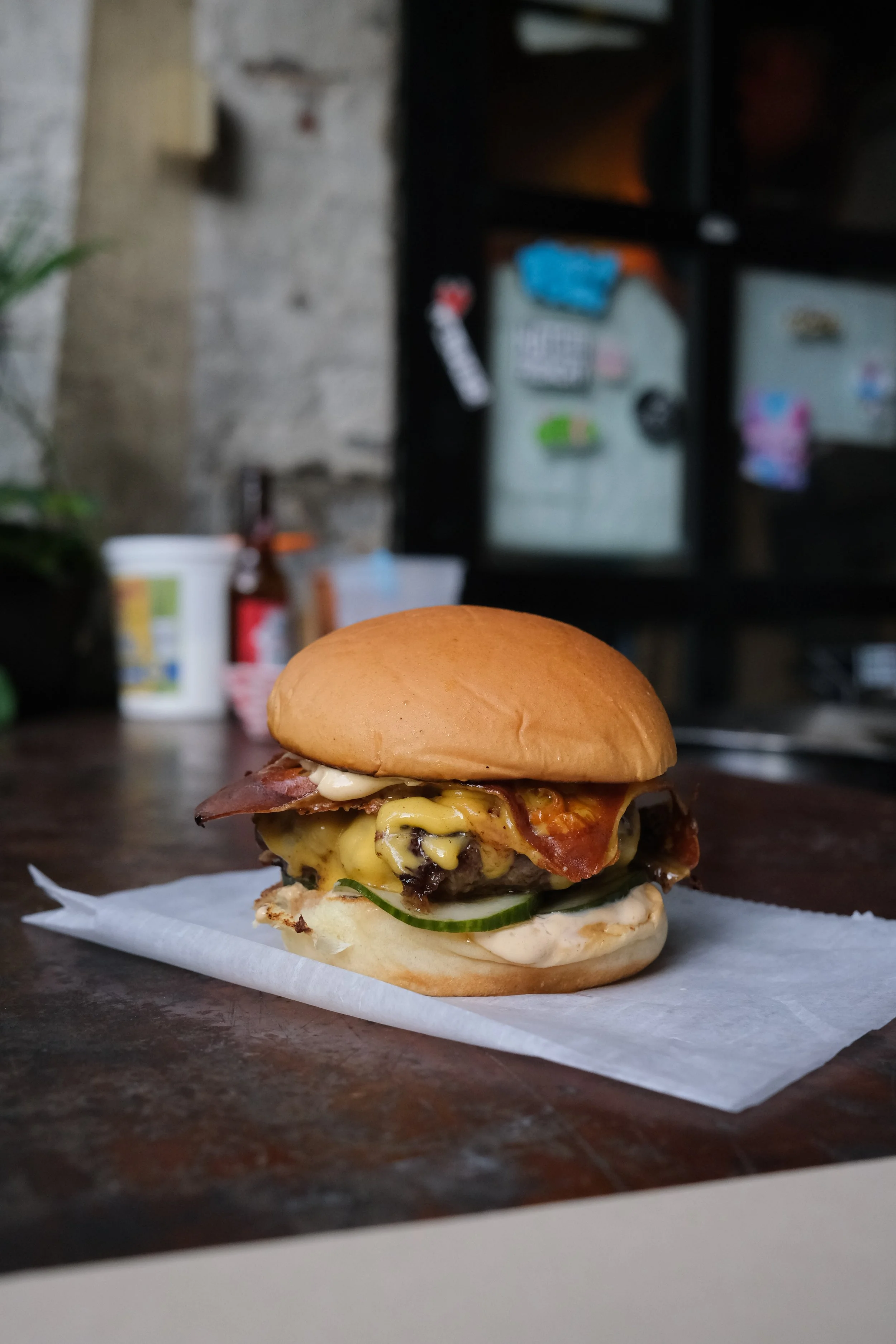 Close-up of a cheeseburger with bacon, pickles, and mayonnaise on a bun, placed on a piece of paper on a wooden surface.