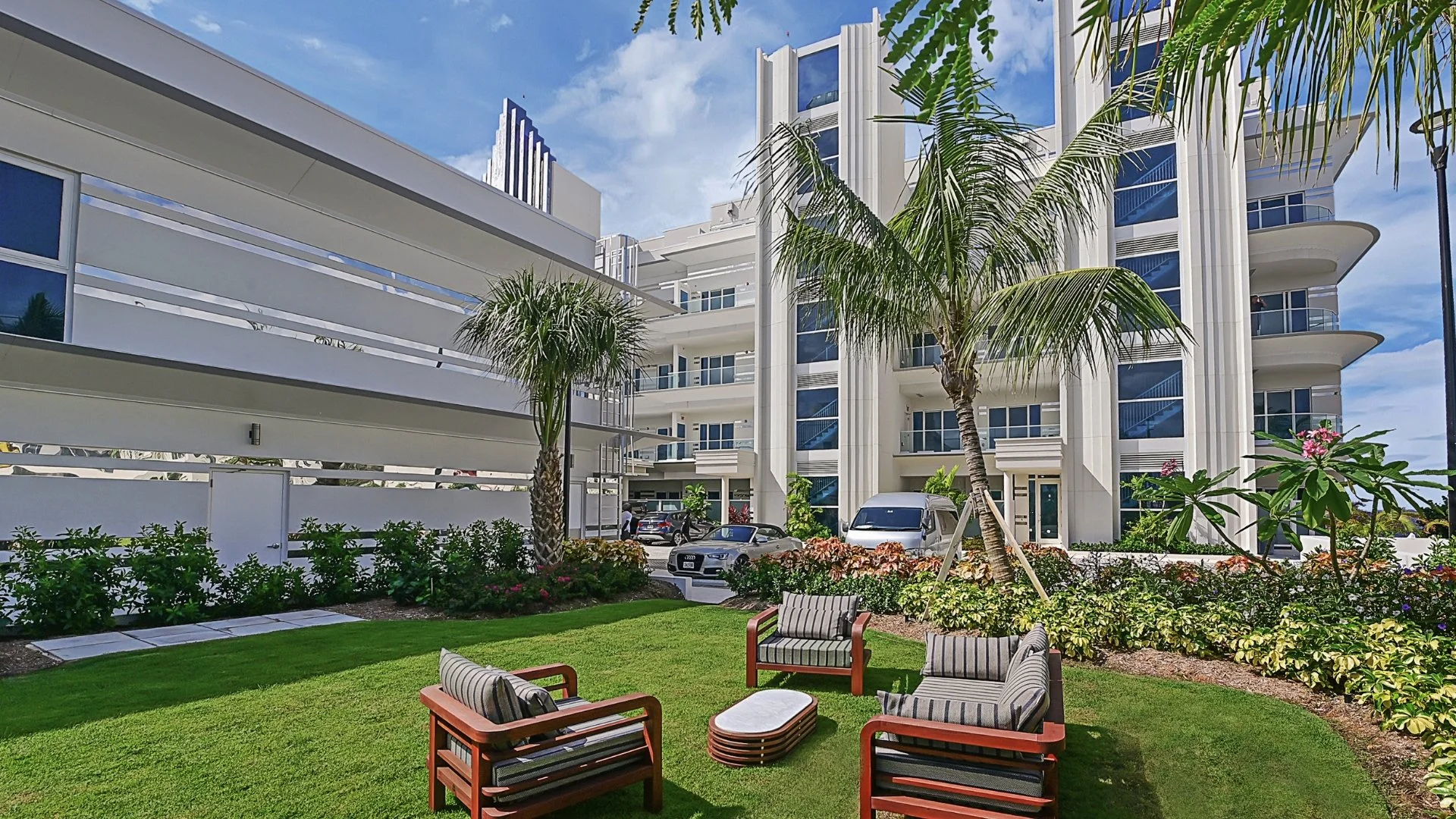 A ground-level view of a manicured green grass courtyard in front of a modern white apartment building with Art Deco style architecture.