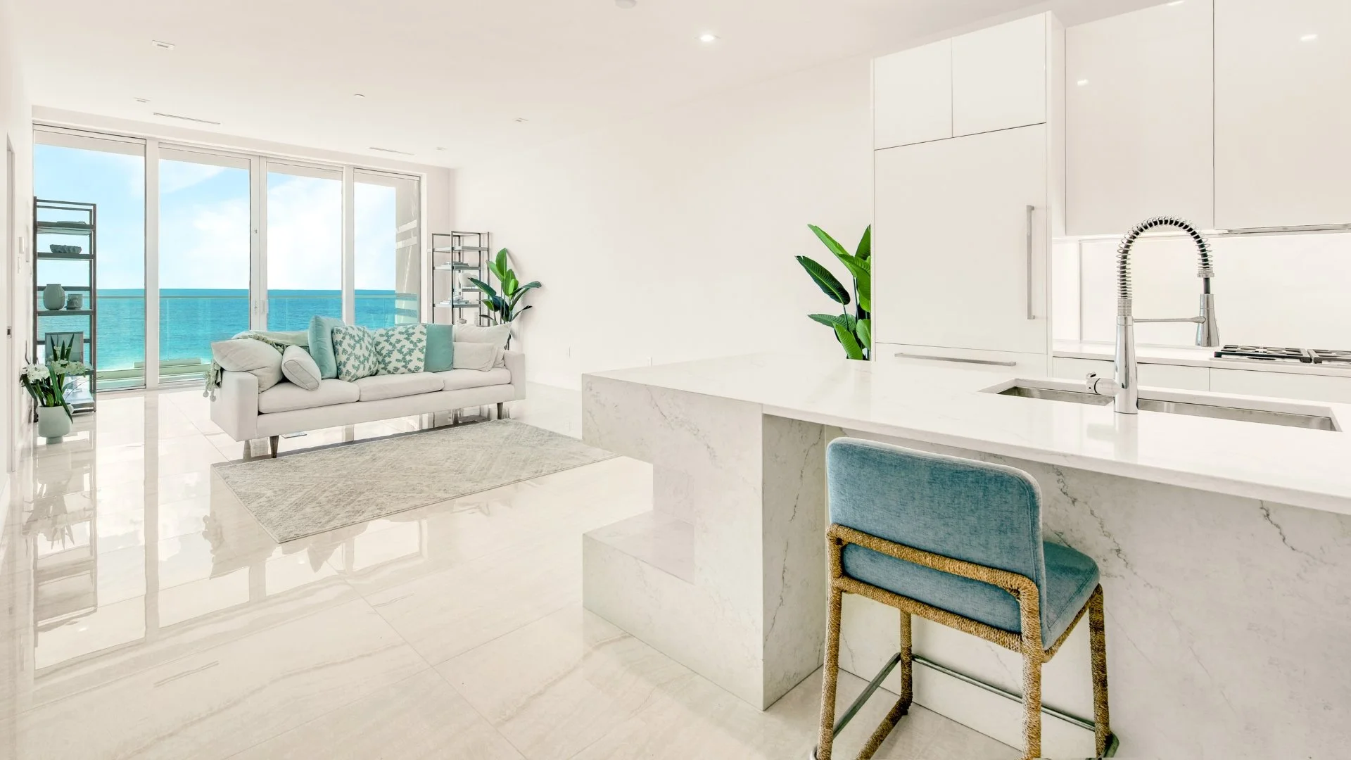 Bright, minimalist interior of a luxury condo showing a white marble kitchen island and a living area with ocean views through glass doors.