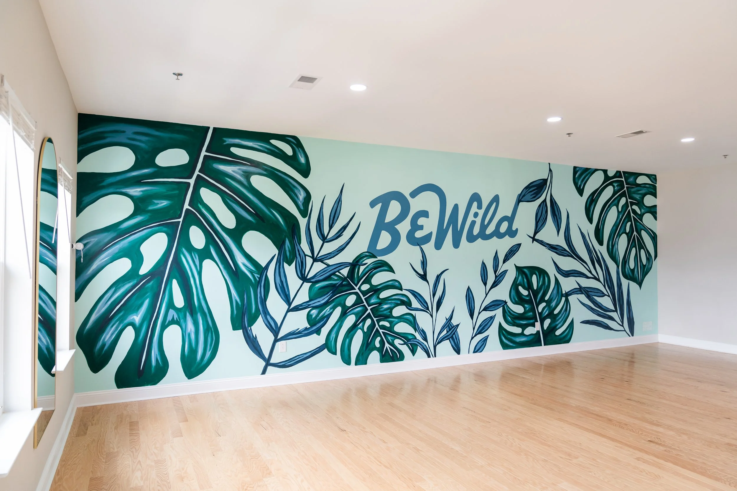 Artistic mural in shades of blue with monstera and palm leaves, highlighting the words 'Be Wild' at the center for Be Wild Yoga studio.