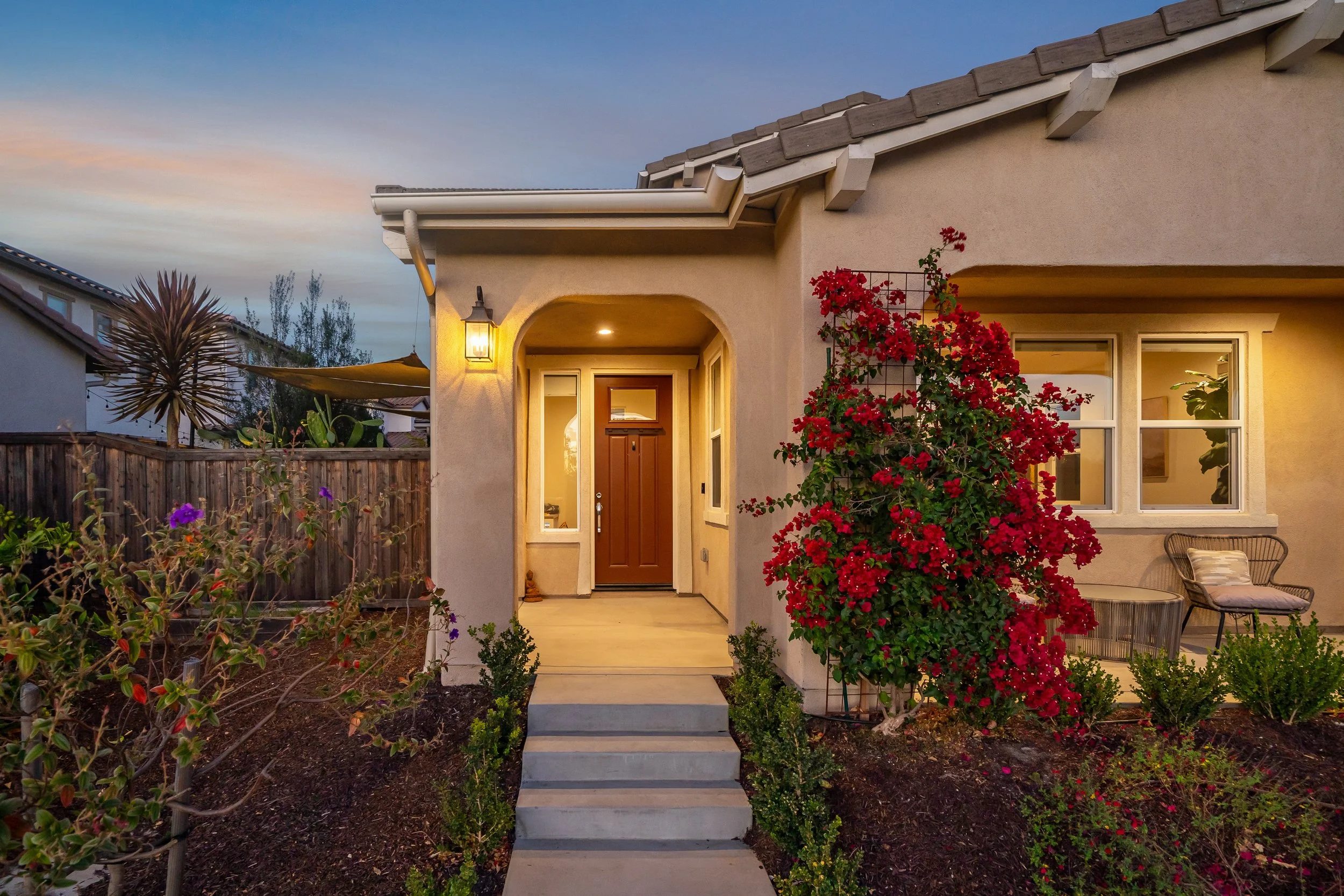 A front entrance of a house with a brown door, outdoor lighting, a porch with chairs, and a garden with flowers and red flowering vine.