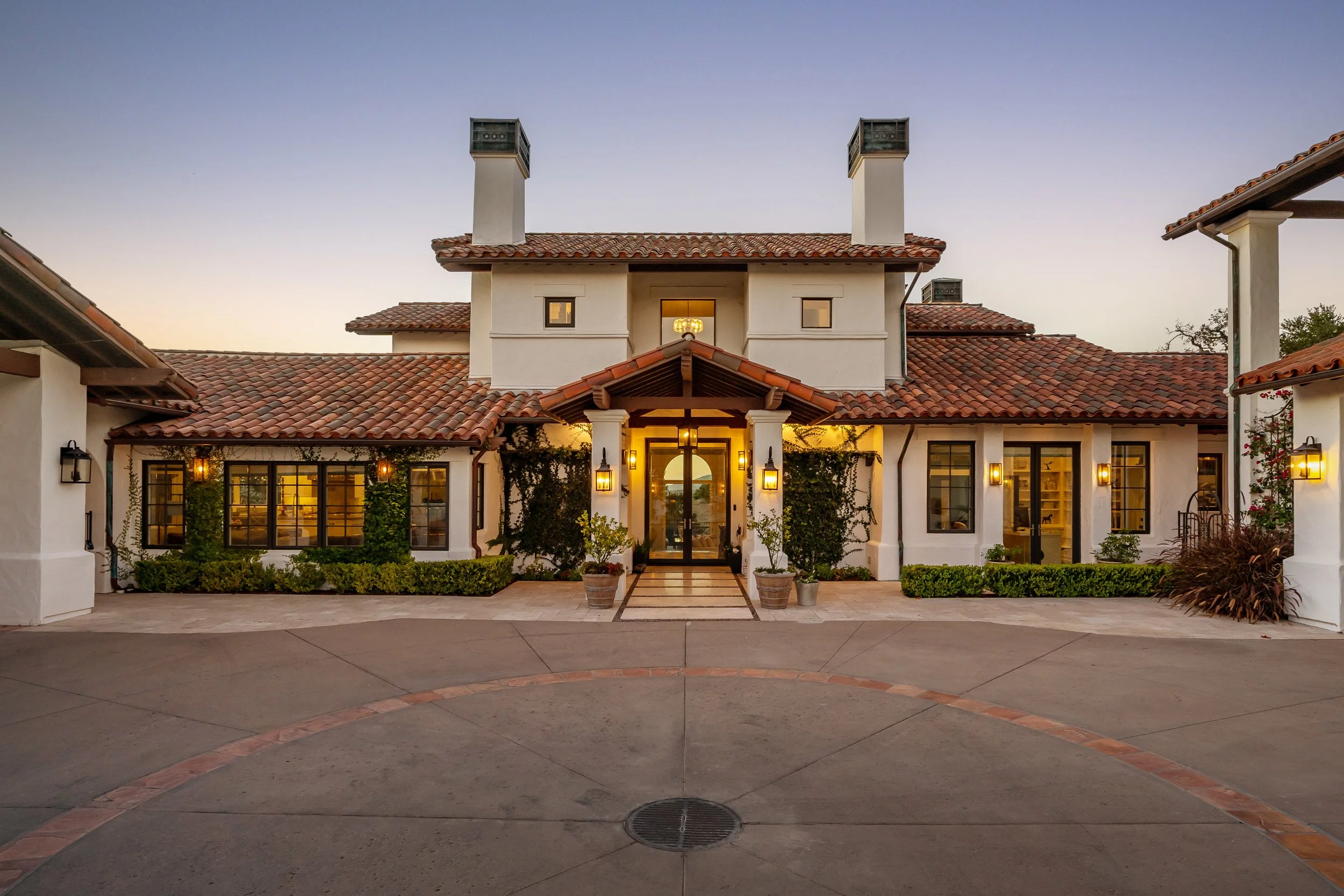 Elegant house with white stucco walls, red tiled roof, multiple chimneys, and warm outdoor lighting, surrounded by landscaped greenery.