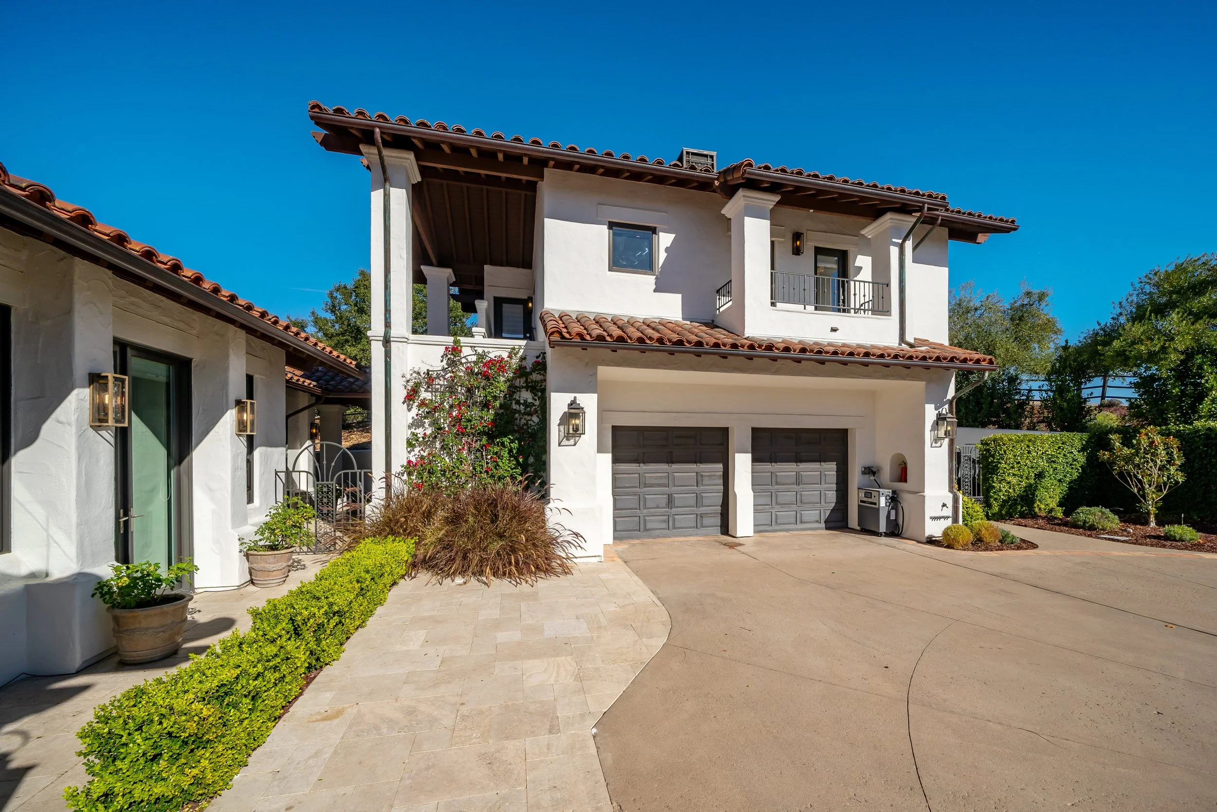 A two-story white house with a red tile roof, black garage doors, and outdoor lighting. The driveway is paved, and there are plants, shrubs, and trees around the house under a clear blue sky.