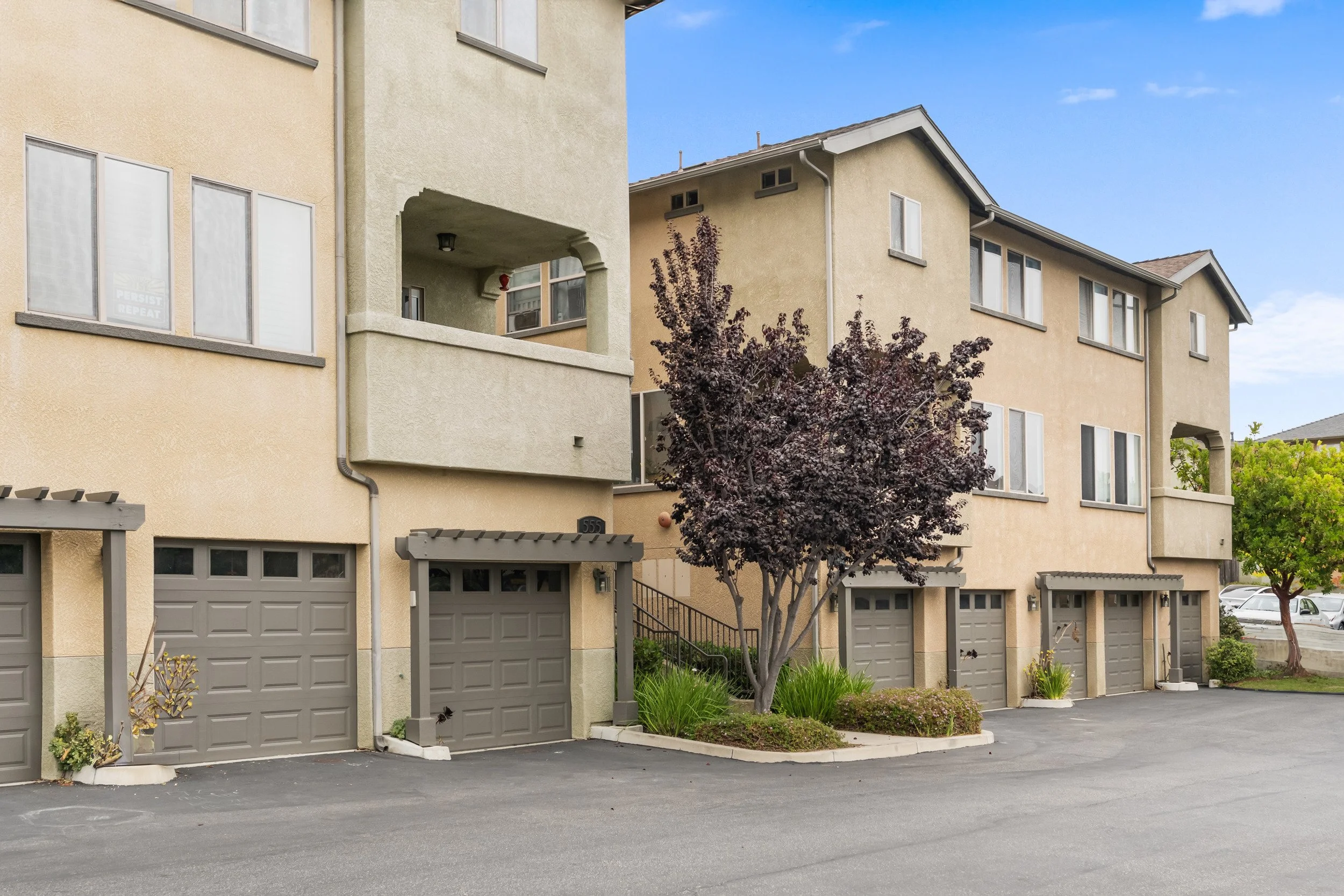 A residential apartment complex with multiple beige buildings, gray garage doors, and landscaped trees and shrubs in the parking lot.