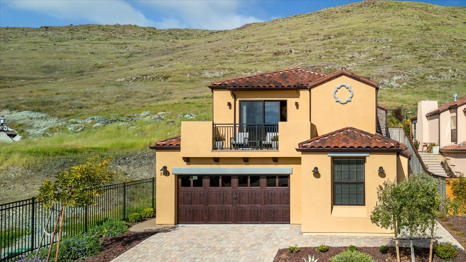 Front view of a two-story house with a beige stucco exterior and a red tiled roof, featuring a garage with a wooden door, a second-floor balcony with potted plants, and landscaped yard with trees and shrubs, set against a grassy hillside background.