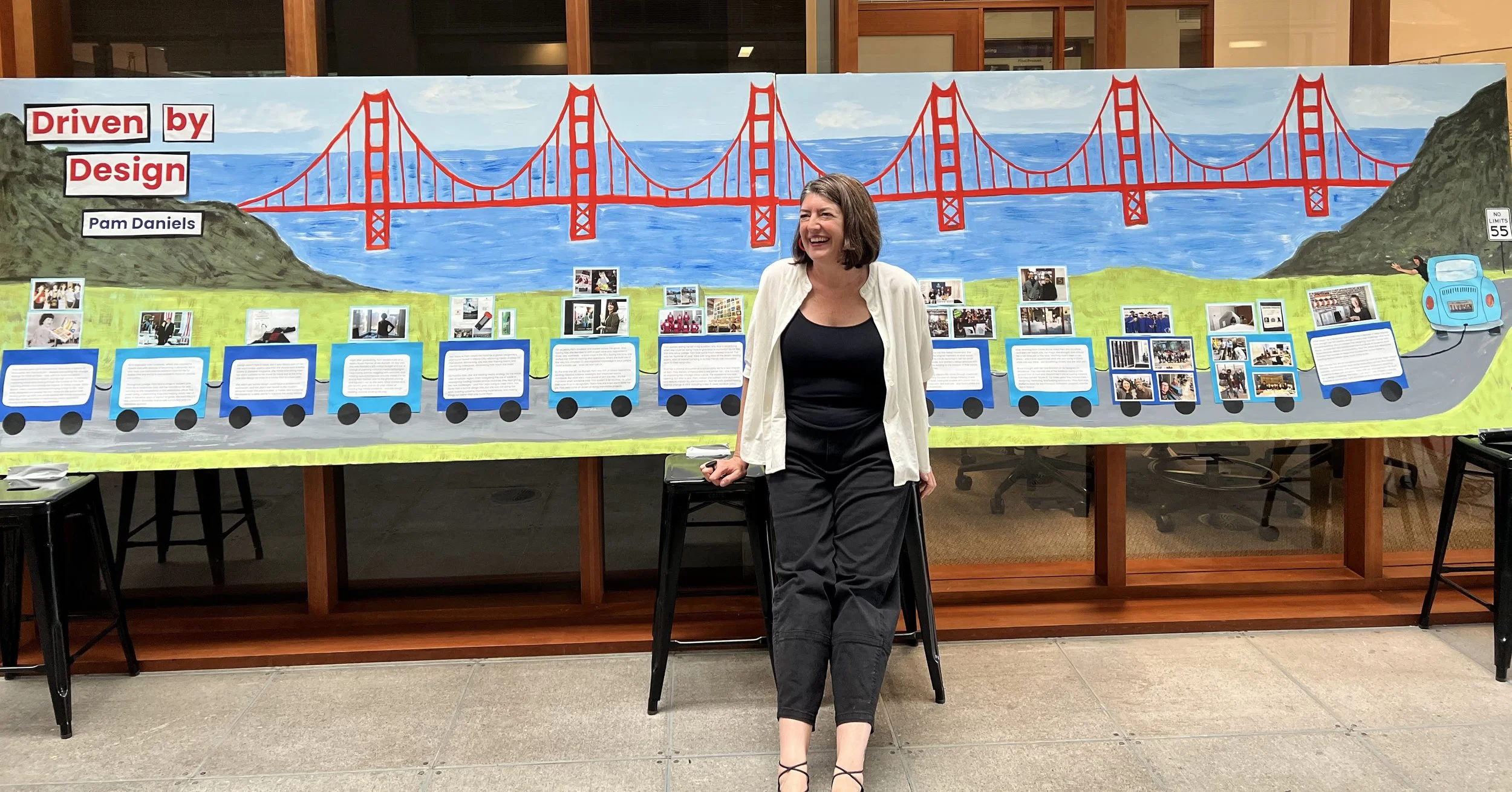 Pam Daniels sitting on a stool in front of a long, colorful collage depicting her career as milestones entitled "Driven by Design" with a backdrop of the Golden Gate Bridge