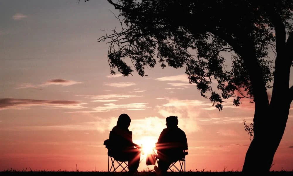 Silhouetted against a vibrant sunset, two people sit on chairs under a tree, sharing a peaceful moment. The sky glows with warm orange and pink hues.