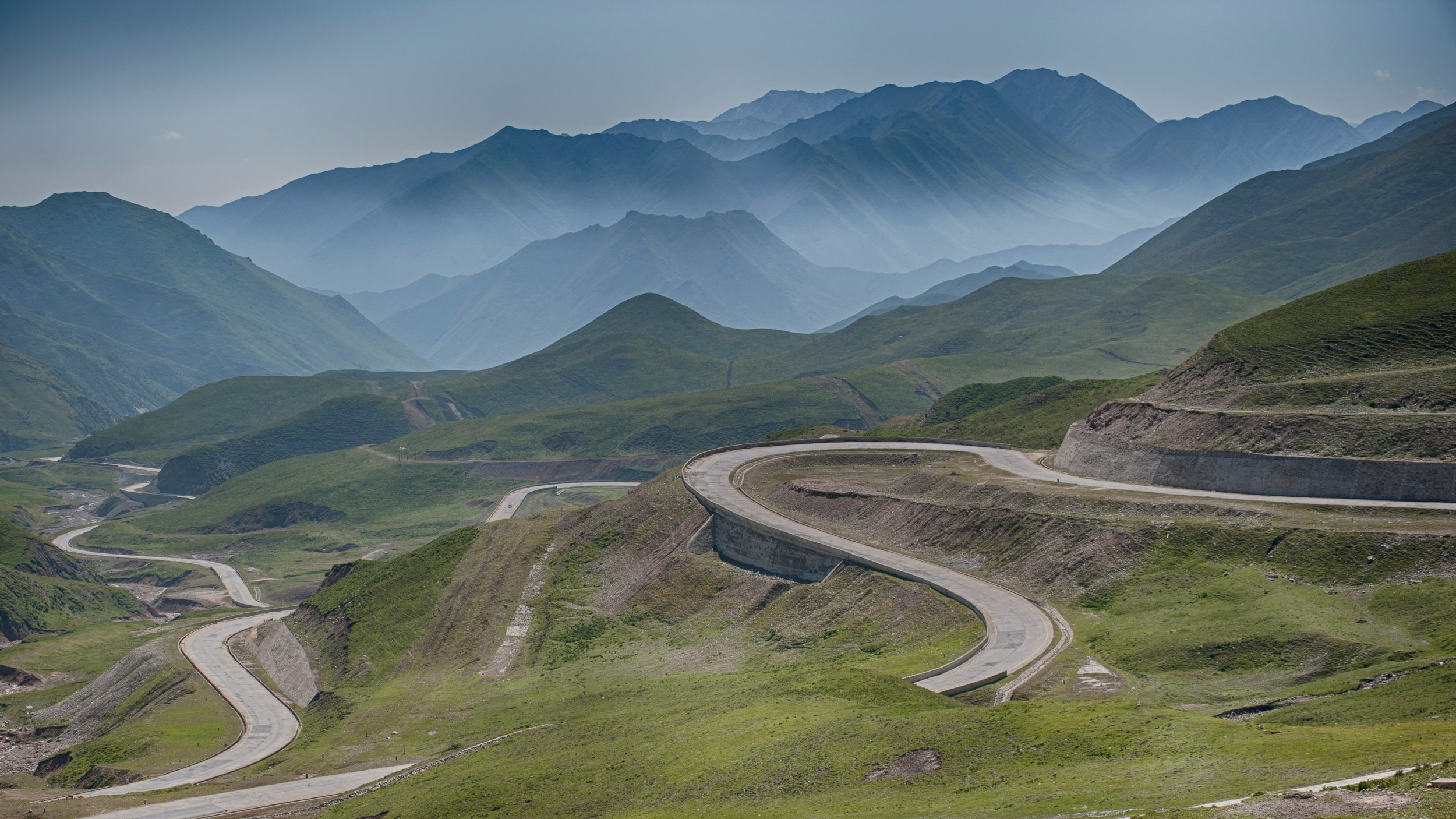 A winding road curves through lush green hills under a hazy blue sky, with distant, misty mountains creating a serene and expansive backdrop.