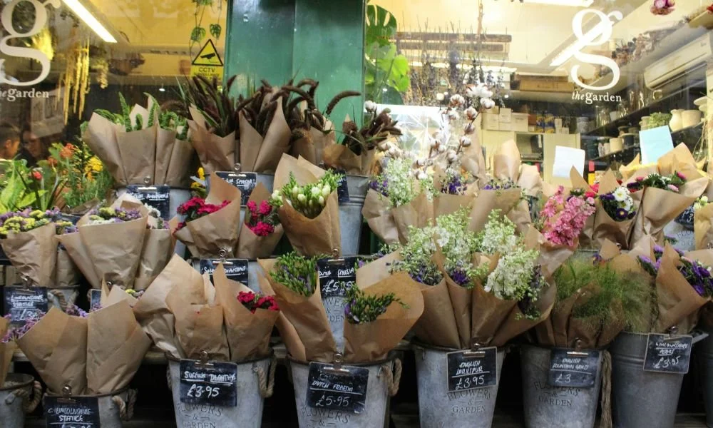 Bouquets of vibrant flowers wrapped in brown paper are displayed in metal buckets outside a shop. Each bouquet is marked with a price tag.