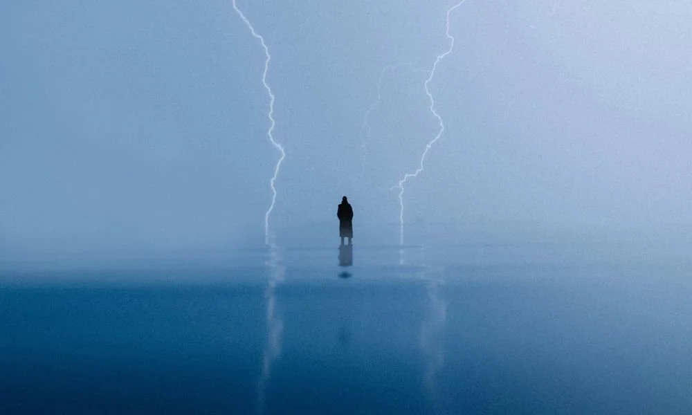A silhouette stands on reflective water under a stormy sky, framed by two striking lightning bolts. The scene conveys solitude and drama.