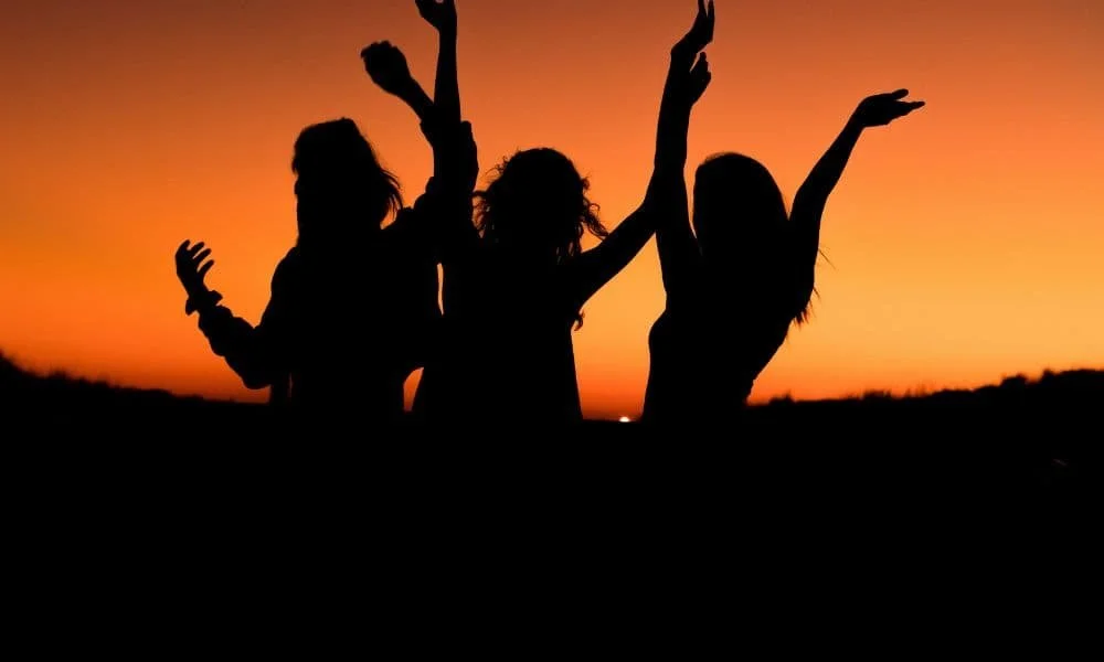 Silhouettes of three people dancing joyfully with raised arms against a vibrant orange sunset sky, conveying celebration and freedom.