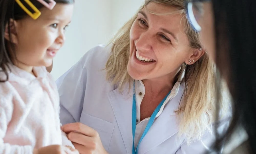 A smiling woman in a white coat, likely a doctor, warmly interacts with a young girl wearing pink. The scene conveys a comforting, joyful atmosphere.