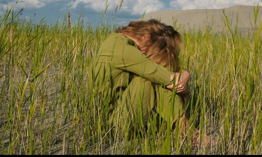 Woman in green clothing sitting in tall grass, hugging knees, with a bowed head. She appears contemplative or sad. Blue sky with clouds in the background.