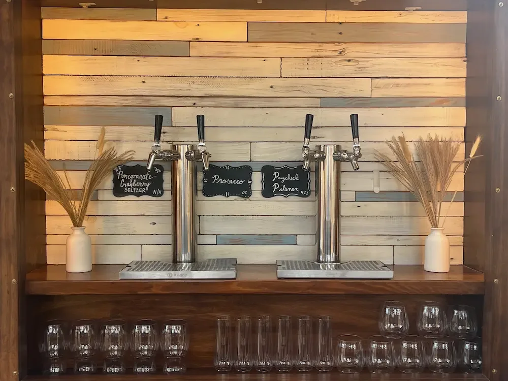 Bar with four taps, black chalkboard signs for each tap, and glasses underneath, flanked by two white vases with dried wheat stalks, against a wood plank wall.